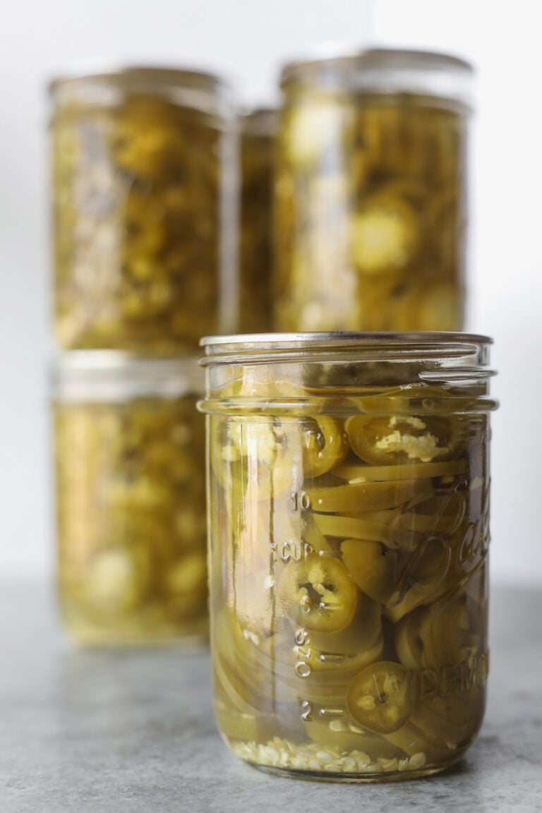 Four Glass Jars Filled With Pickled Jalapeños Are Arranged On A Gray Surface, With One Jar In The Front And Three Jars Clustered Together In The Background.