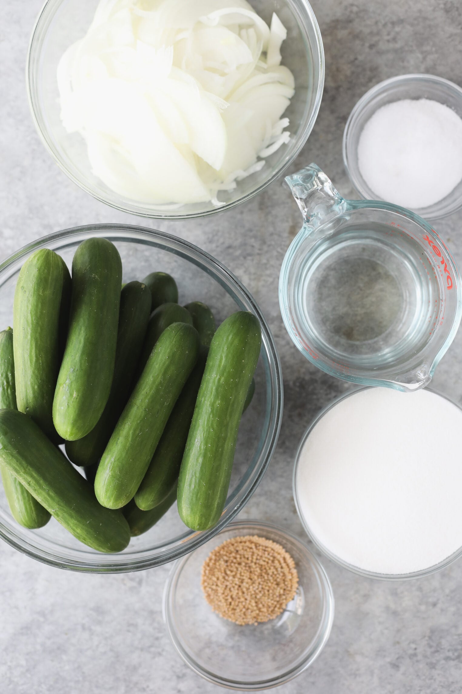 Bowls Of Sliced Onions, Mini Cucumbers, Salt, Mustard Seeds, And Sugar—Classic Ingredients For Bread And Butter Pickles—Plus A Measuring Cup Of Water, Arranged On A Grey Surface Viewed From Above.