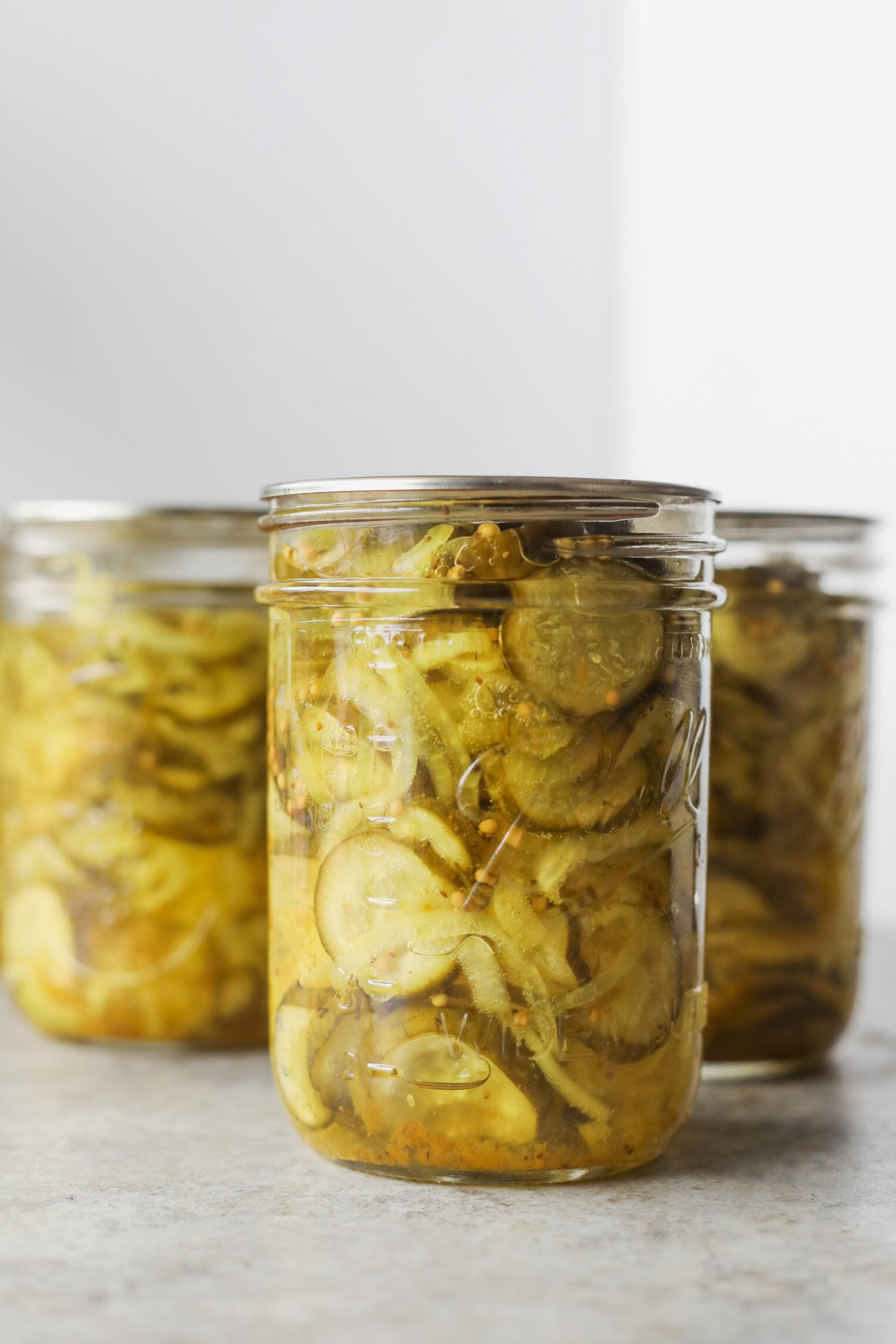Three Glass Jars Filled With Homemade Bread And Butter Pickles, Showing Sliced Cucumbers And Onions In A Yellow Brine, Are Arranged On A Light Surface With A White Background.