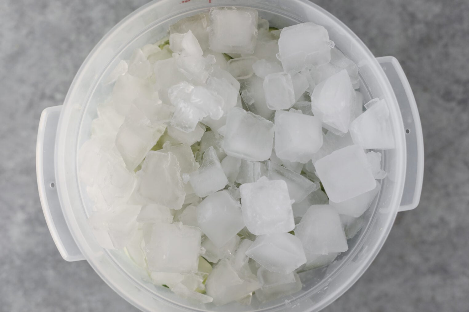 A Clear Round Container Filled With Small Ice Cubes And A Generous Helping Of Bread And Butter Pickles Sits On A Gray Surface, Viewed From Above.