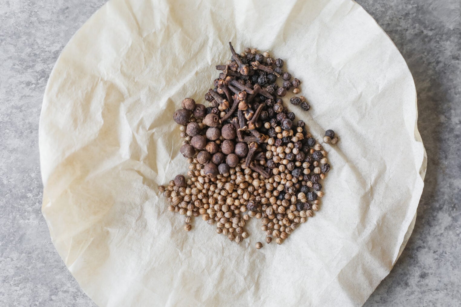 A Pile Of Whole Spices, Including Coriander Seeds, Black Peppercorns, Allspice, And Cloves, Sits On A Piece Of Crumpled Parchment Paper On A Gray Surface—Perfect Ingredients For Making Your Own Bread And Butter Pickles.