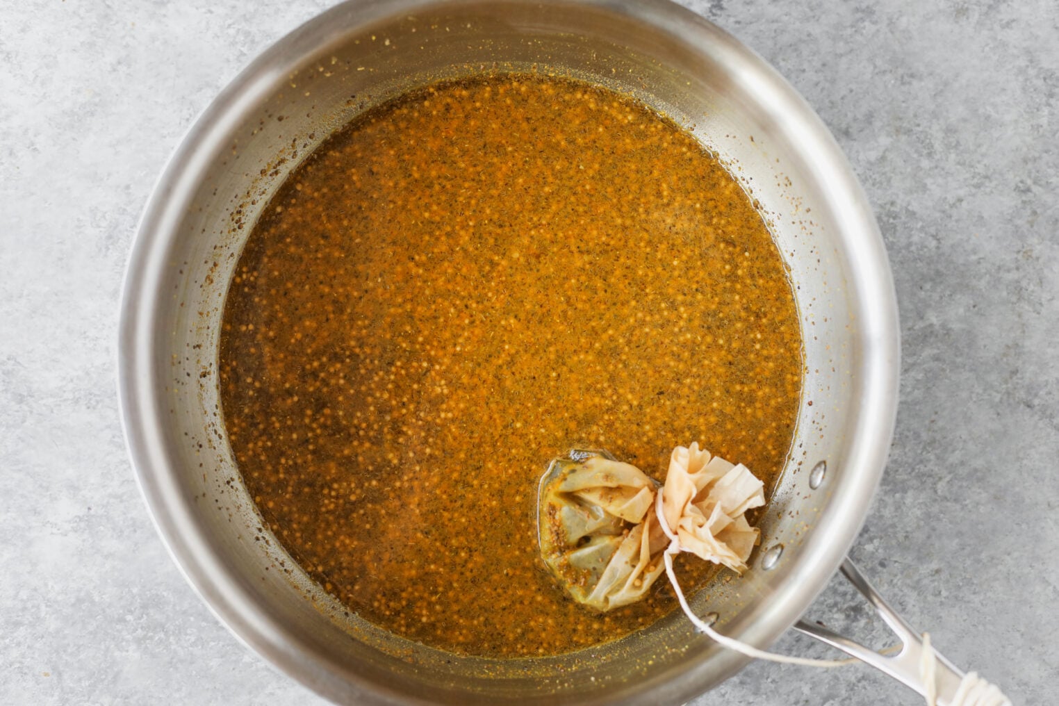 A Metal Pot Filled With A Golden-Brown Spiced Liquid, Reminiscent Of The Brine Used For Bread And Butter Pickles, With A Used Tea Bag Resting On The Surface And Tied To The Pot Handle. The Background Is A Light Gray Countertop.