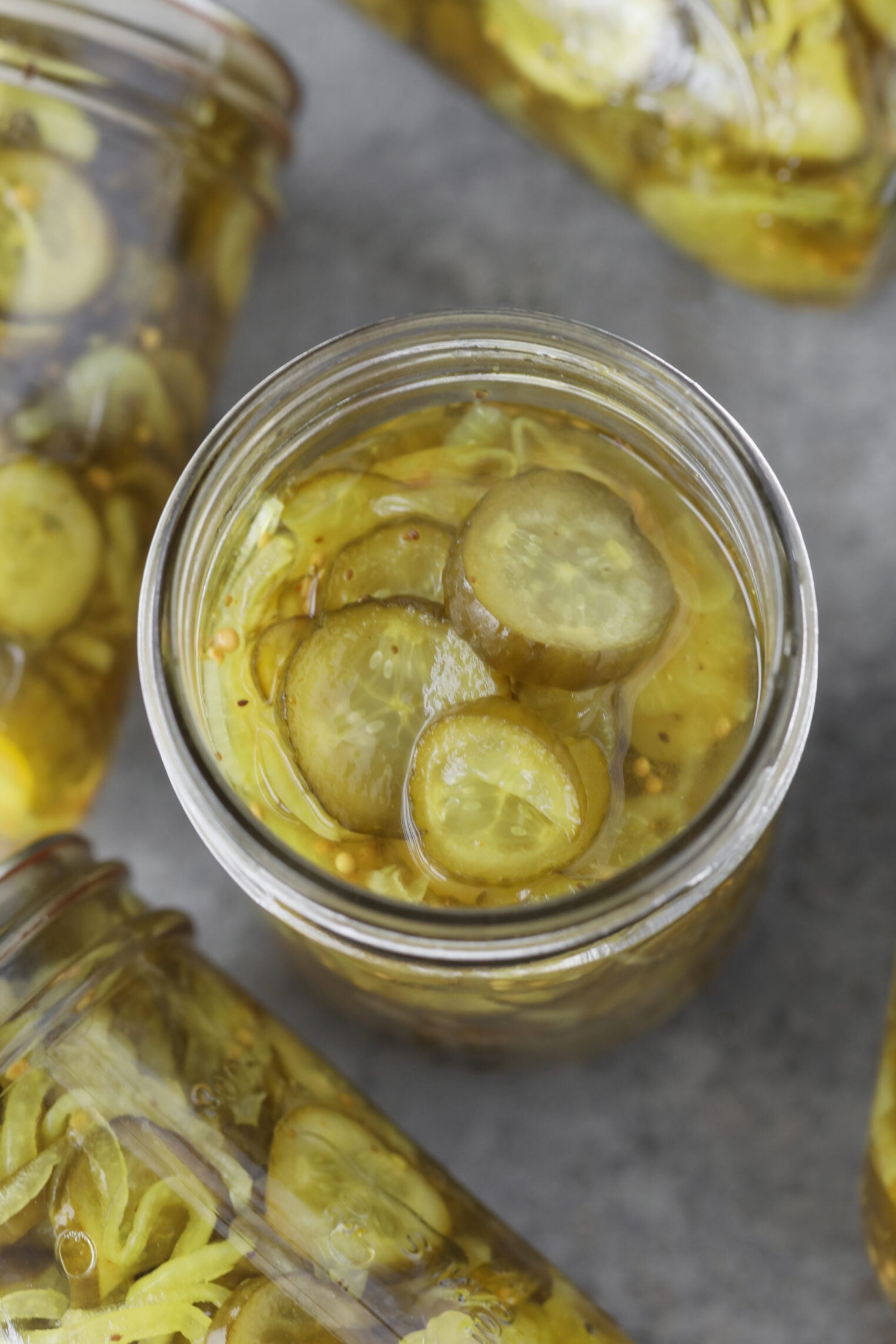 A Glass Jar Filled With Sliced Bread And Butter Pickles In Brine, Surrounded By Other Similar Jars On A Gray Surface.