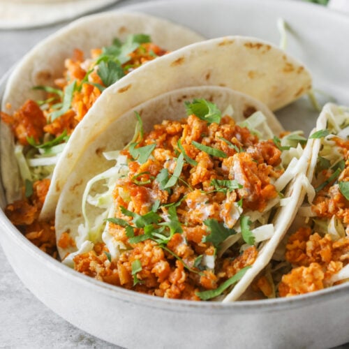 Three Soft Tacos Filled With Flavorful Buffalo Tofu, Shredded Cabbage, And Fresh Cilantro Are Served In A White Bowl. Flour Tortillas And Cilantro Are Visible In The Background.