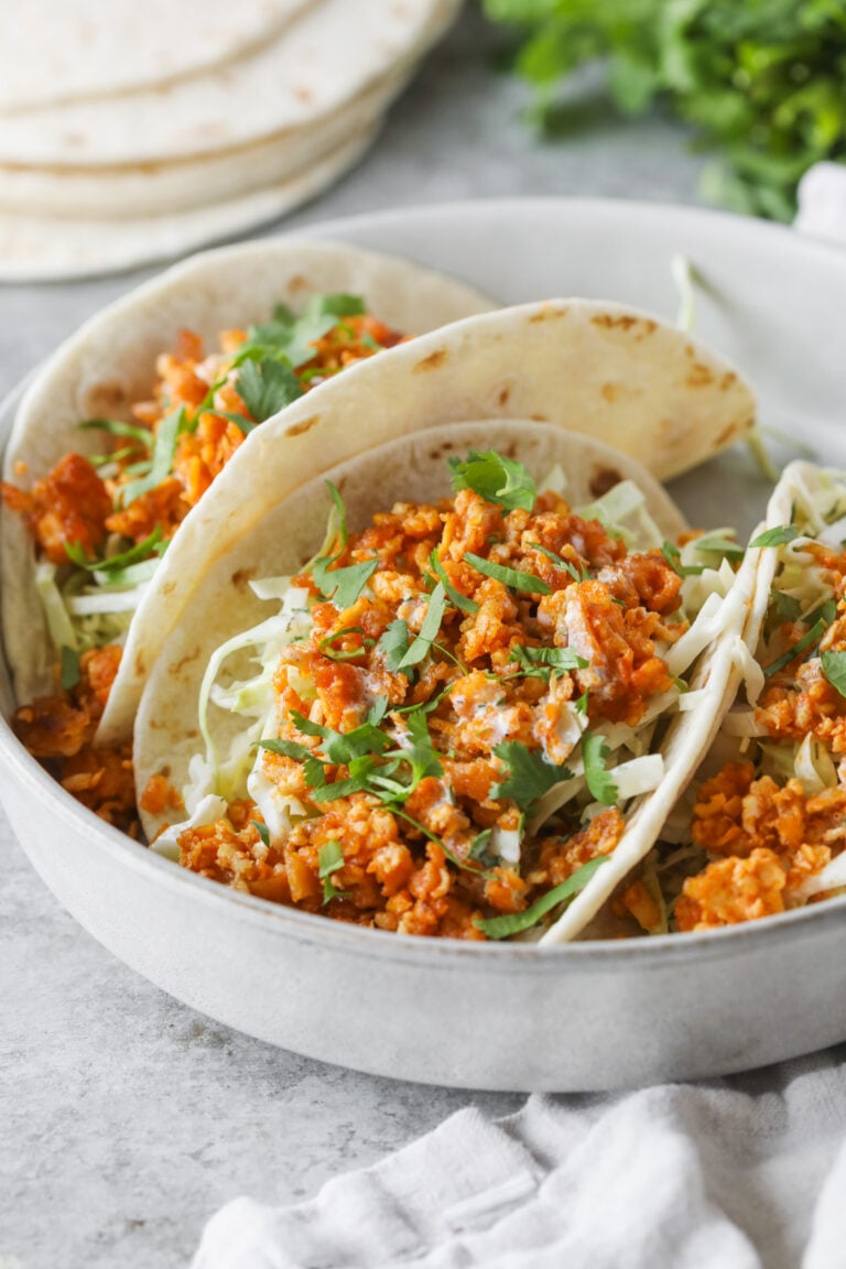 Three Soft Tacos Filled With Flavorful Buffalo Tofu, Shredded Cabbage, And Fresh Cilantro Are Served In A White Bowl. Flour Tortillas And Cilantro Are Visible In The Background.