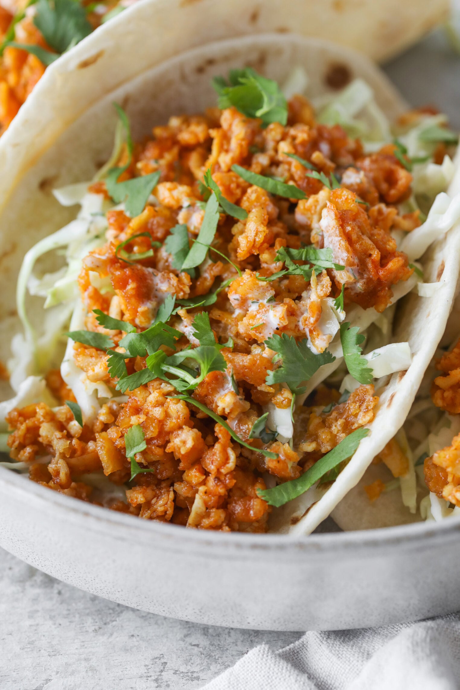 A Close-Up Of Buffalo Tofu Tacos Filled With Seasoned Ground Meat, Shredded Lettuce Or Cabbage, Creamy Sauce, And Fresh Cilantro, Served In A Light-Colored Bowl.