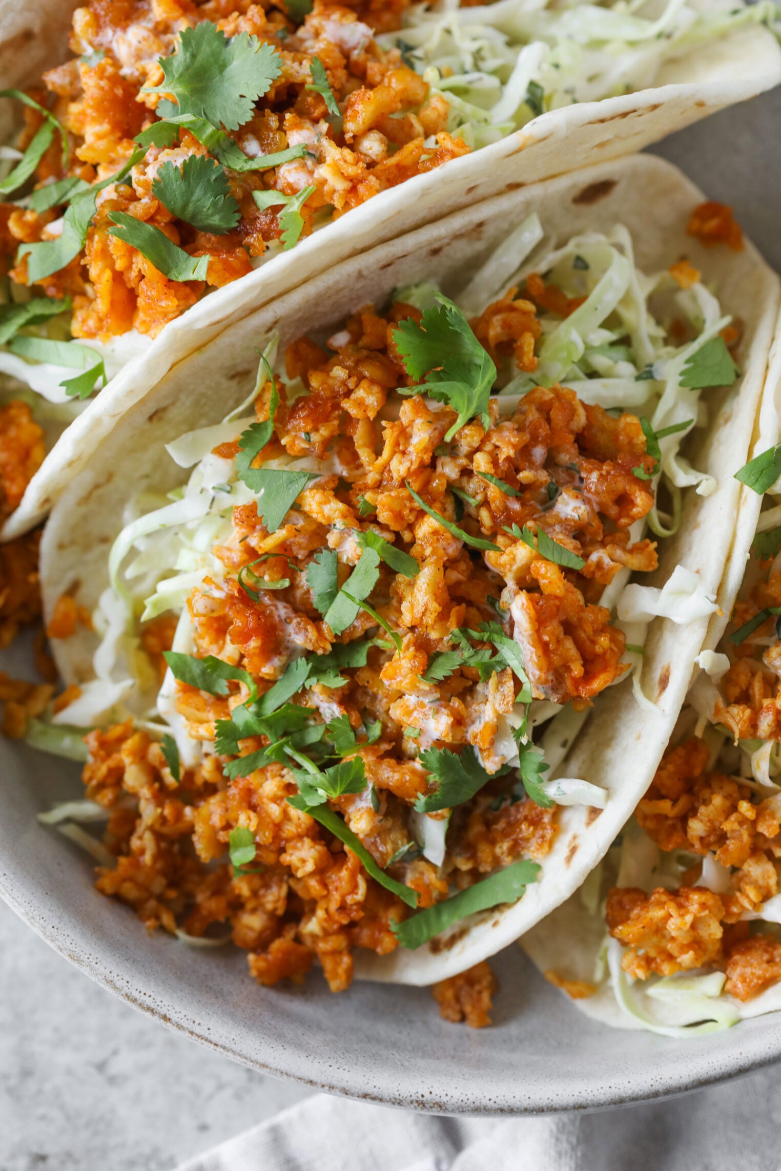 Three Soft Tacos Filled With Seasoned Buffalo Tofu Tacos Crumbles, Shredded Cabbage, And Topped With Chopped Cilantro, Served On A Light-Colored Plate.