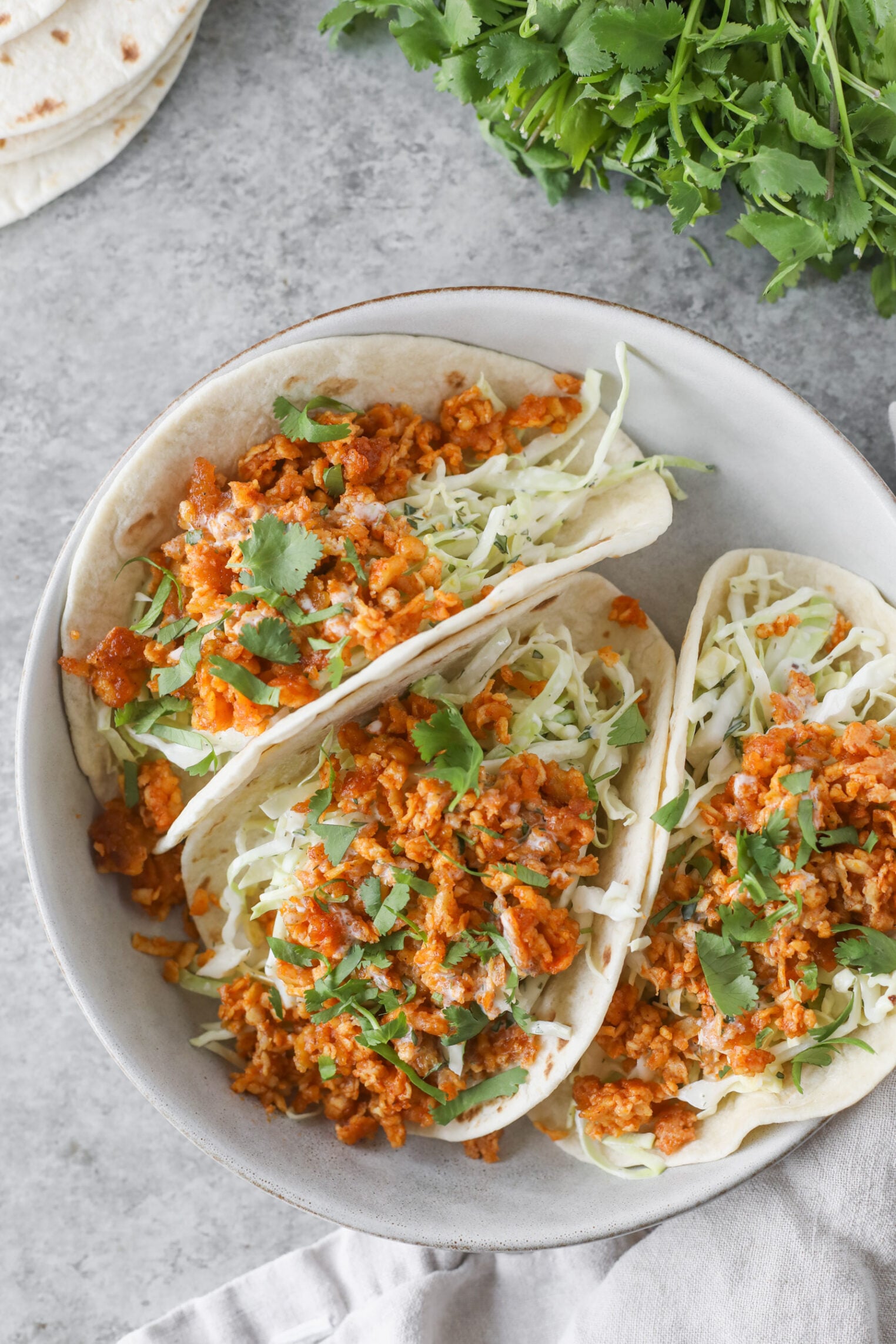 Three Soft Buffalo Tofu Tacos Filled With Shredded Cabbage, Seasoned Crumbled Tofu, And Chopped Cilantro Are Arranged In A Bowl, With Fresh Cilantro And Tortillas On The Side.