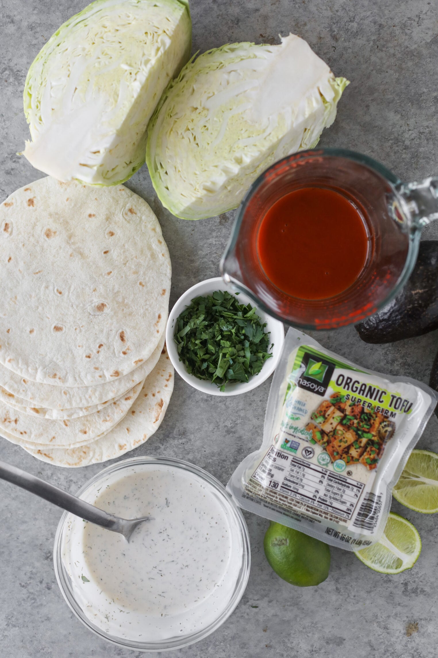 A Flat Lay Of Taco Ingredients For Buffalo Tofu Tacos On A Gray Surface: Sliced Cabbage, Flour Tortillas, Chopped Cilantro, A Glass Measuring Cup Of Red Sauce, Organic Tofu, Creamy Sauce With A Spoon, Halved Lime, And Avocado.