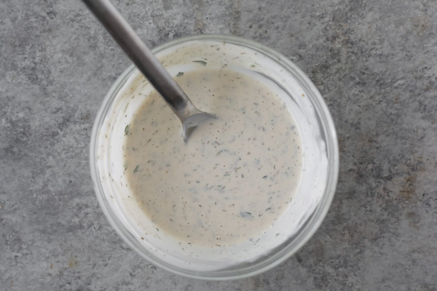 A Glass Bowl Filled With Creamy Ranch Dressing Dotted With Herbs, With A Metal Spoon Resting Inside. Perfect For Drizzling Over Buffalo Tofu Tacos, The Bowl Sits On A Gray, Textured Surface.