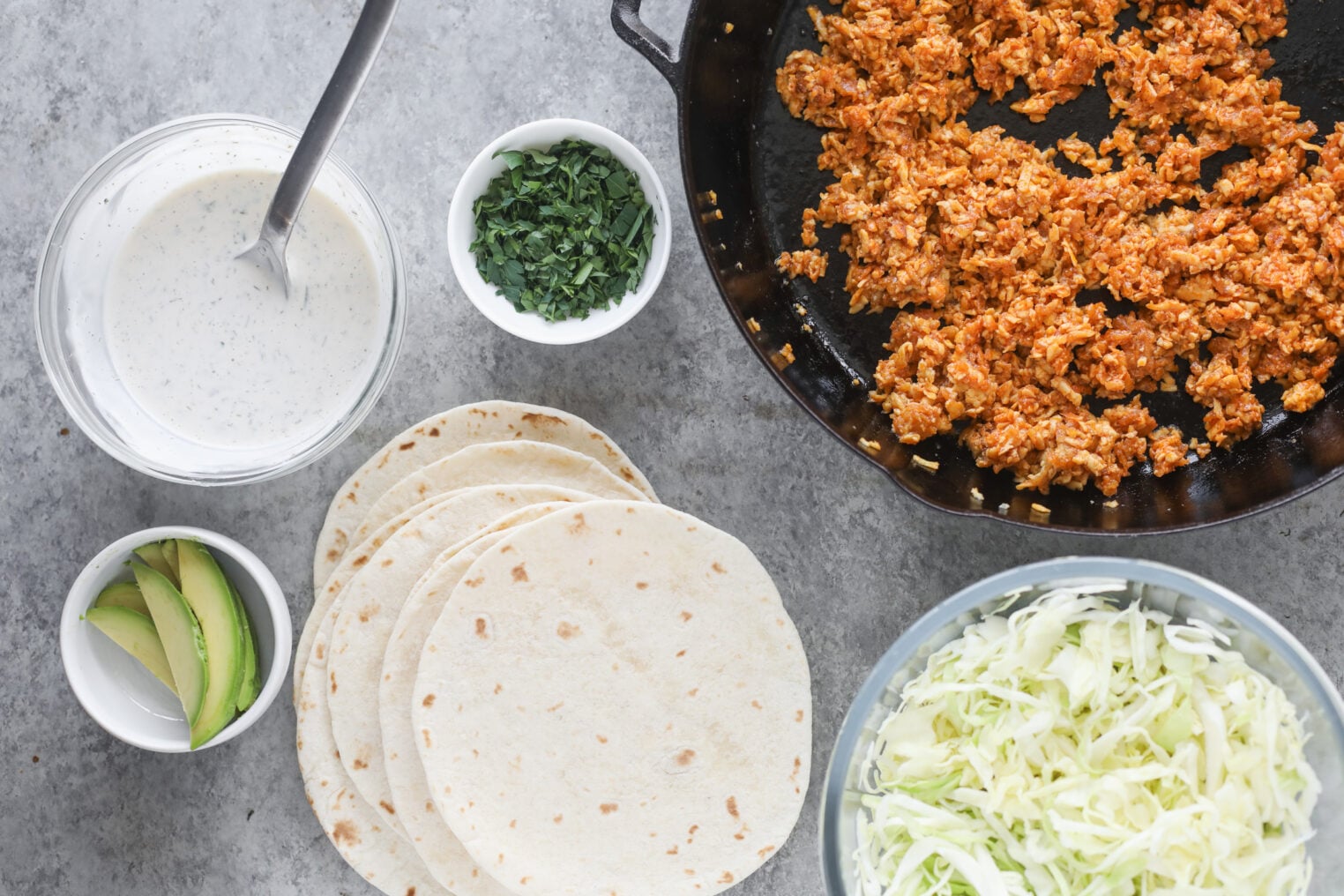 Ingredients For Buffalo Tofu Tacos Laid Out On A Gray Surface: A Skillet With Seasoned Crumbled Filling, Tortillas, Shredded Cabbage, Avocado Slices, Creamy Sauce In A Bowl With A Spoon, And Chopped Herbs In A Small Bowl.