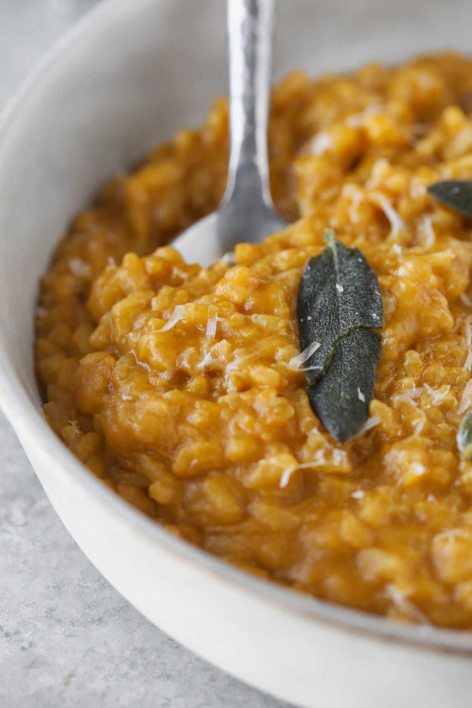 Butternut Squash Risotto 7 A Close-Up Of Creamy Butternut Squash Risotto Topped With Grated Cheese And Crispy Sage Leaves, Served In A White Bowl With A Metal Spoon.