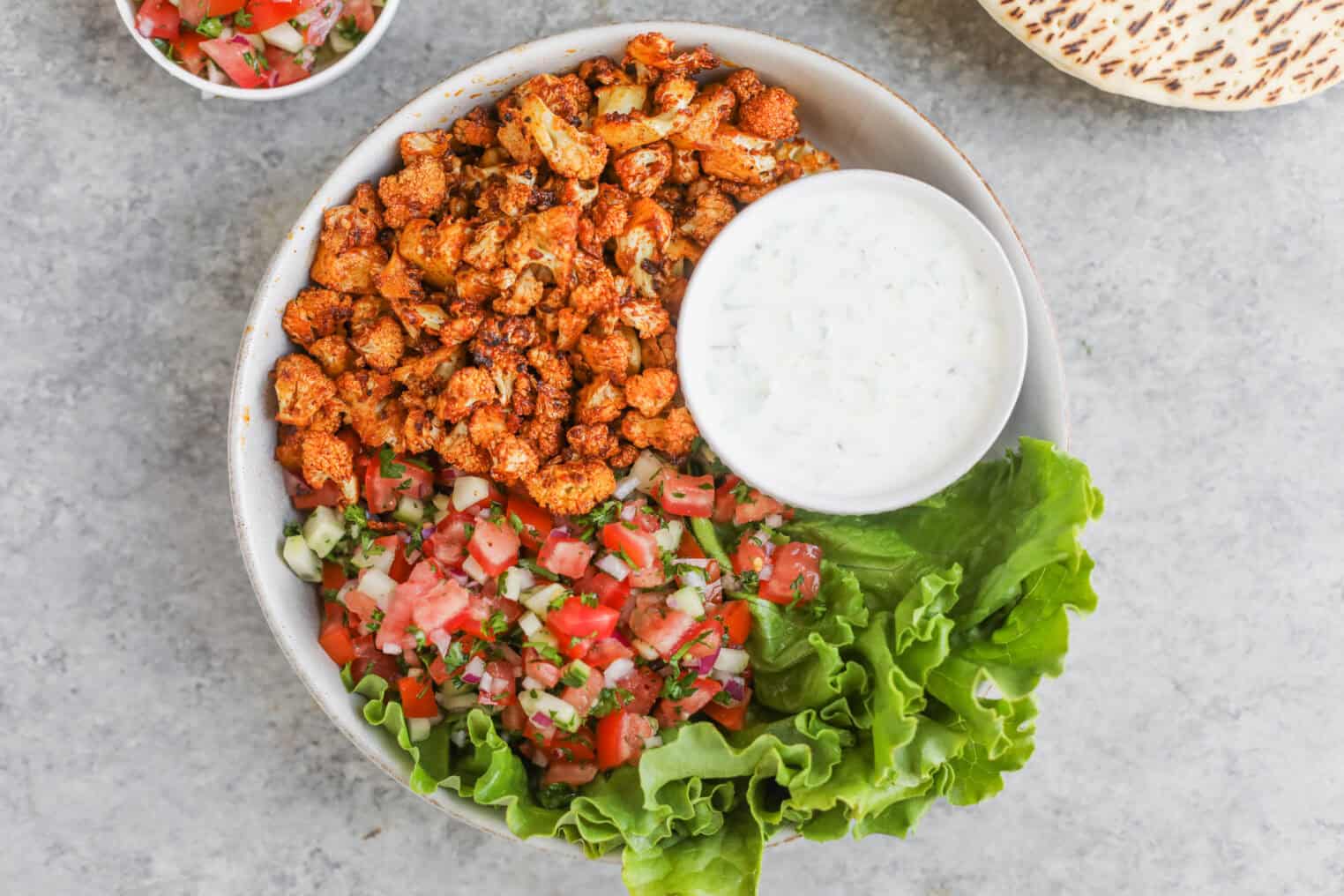 Cauliflower Shawarma 7 A Bowl Containing Spiced Grilled Chicken Pieces, Cauliflower Shawarma, Fresh Lettuce Leaves, Diced Tomato And Cucumber Salad, And A Small Bowl Of Creamy White Sauce, All Arranged On A Gray Surface.