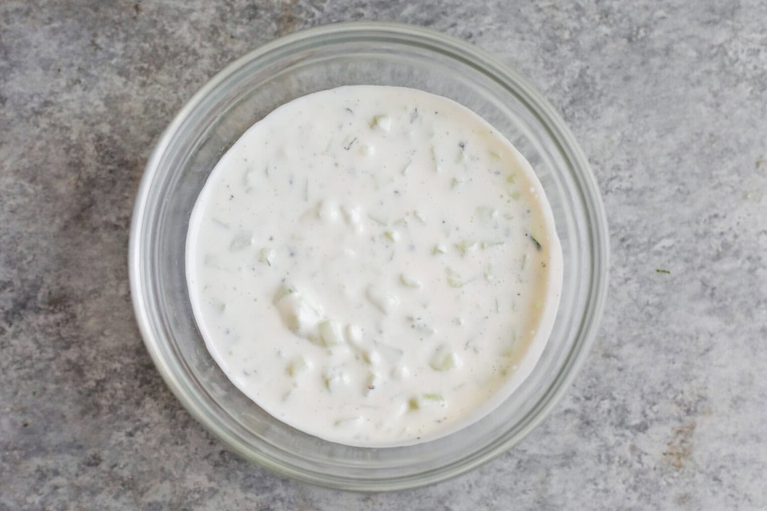 Cauliflower Shawarma 5 A Glass Bowl Filled With Creamy White Tzatziki Sauce, Containing Small Pieces Of Cucumber And Herbs, Sits On A Gray Textured Surface—Perfect For Serving Alongside Cauliflower Shawarma.