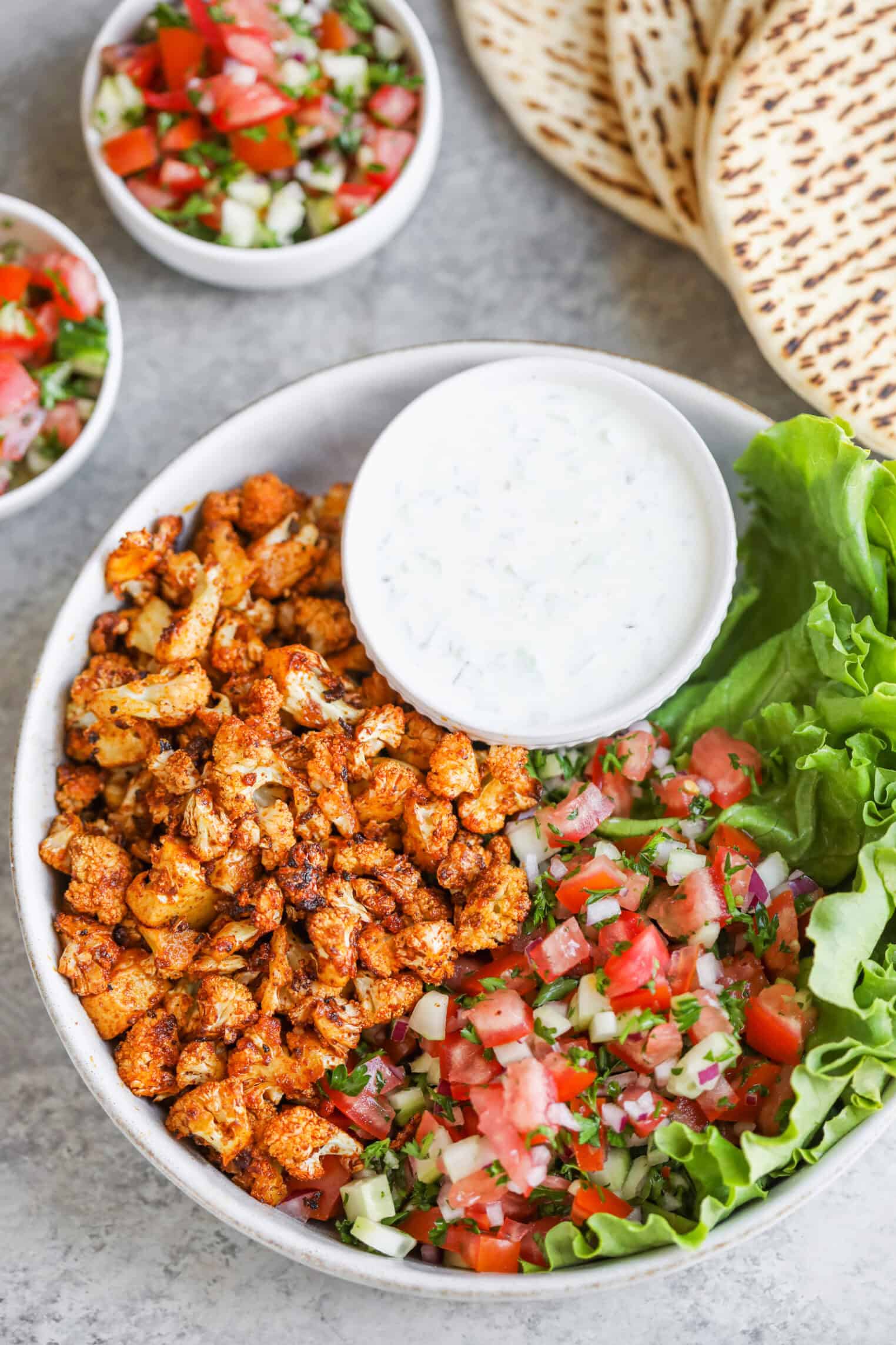 Cauliflower Shawarma 1 A Bowl With Roasted Spiced Cauliflower Shawarma, Fresh Tomato-Cucumber Salad, Leafy Lettuce, And A Small Bowl Of Creamy White Sauce. Pita Bread And Extra Salad Are In The Background.