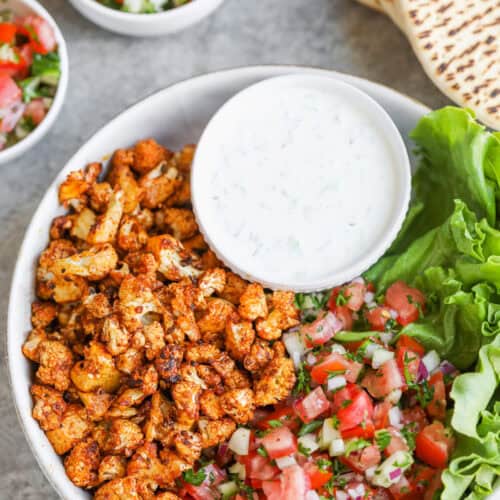Cauliflower Shawarma 11 A Bowl With Roasted Spiced Cauliflower Shawarma, Fresh Tomato-Cucumber Salad, Leafy Lettuce, And A Small Bowl Of Creamy White Sauce. Pita Bread And Extra Salad Are In The Background.