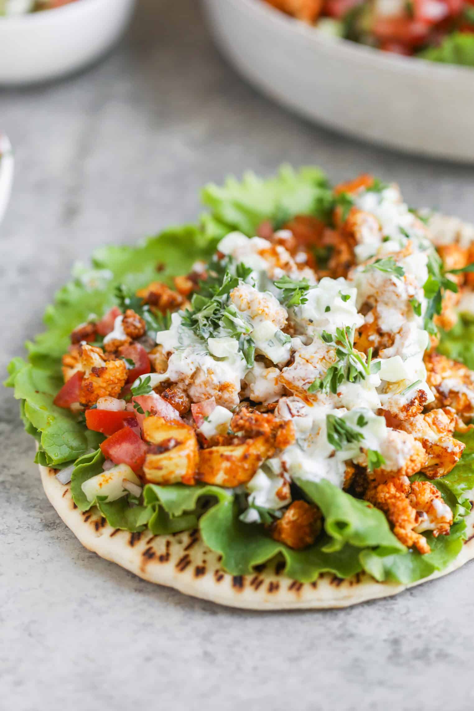 Cauliflower Shawarma 3 A Pita Topped With Fresh Lettuce, Seasoned Grilled Chicken Pieces Or Cauliflower Shawarma, Diced Vegetables, And A Drizzle Of Creamy White Sauce, Garnished With Herbs, On A Light Gray Surface.