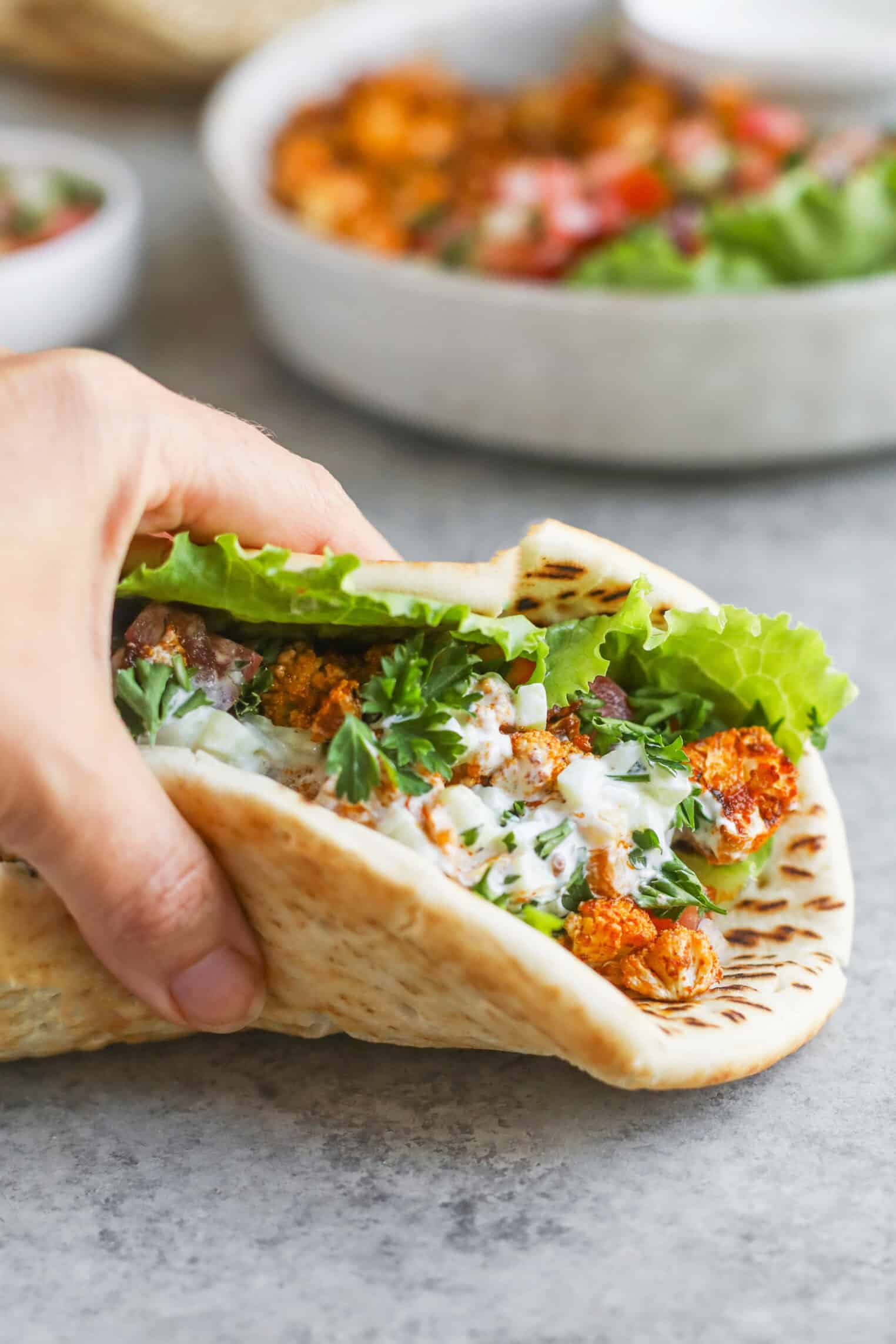 Cauliflower Shawarma 8 A Hand Holds A Pita Wrap Filled With Lettuce, Cauliflower Shawarma, Fresh Herbs, And A Drizzle Of White Sauce On A Gray Surface. A Bowl Of Salad Is Blurred In The Background.