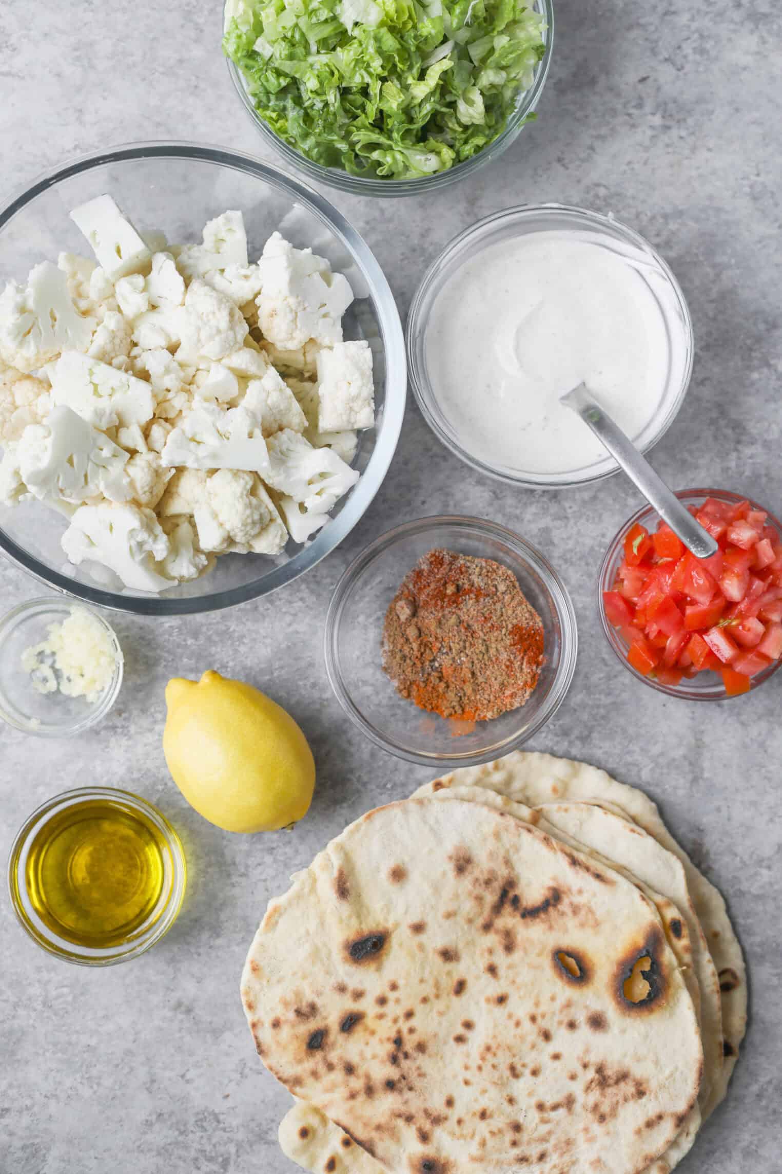 Cauliflower Shawarma 2 A Flatlay Of Ingredients On A Gray Surface: Naan Bread, Lemon, Olive Oil, Minced Garlic, Chopped Lettuce, Cauliflower Florets For Cauliflower Shawarma, Sliced Tomatoes, A Bowl Of Creamy Sauce, A Spice Mix, And A Stack Of Flatbread.