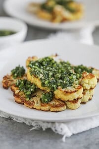 Roasted Cauliflower Steaks Topped With Vibrant Chimichurri Sauce Are Served On A White Plate. The Dish Is Elegantly Presented On A Grey Surface, Accompanied By A Bowl Of Extra Sauce In The Background.
