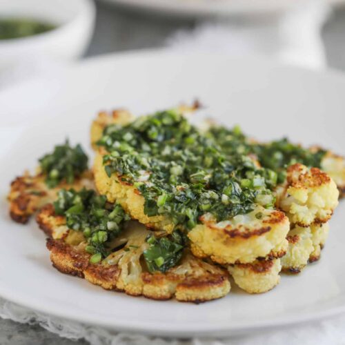 Roasted Cauliflower Steaks Topped With Vibrant Chimichurri Sauce Are Served On A White Plate. The Dish Is Elegantly Presented On A Grey Surface, Accompanied By A Bowl Of Extra Sauce In The Background.