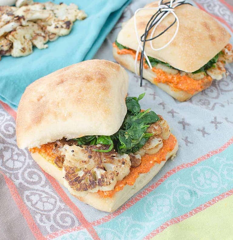 Two Sandwiches On A Colorful Cloth Showcase Healthy Vegan Cookout Recipes. Each Features Roasted Cauliflower, Spinach, And Red Pepper Spread In Ciabatta Buns. One Sandwich Is Tied With String, And Some Roasted Cauliflower Rests On A Blue Napkin Nearby.