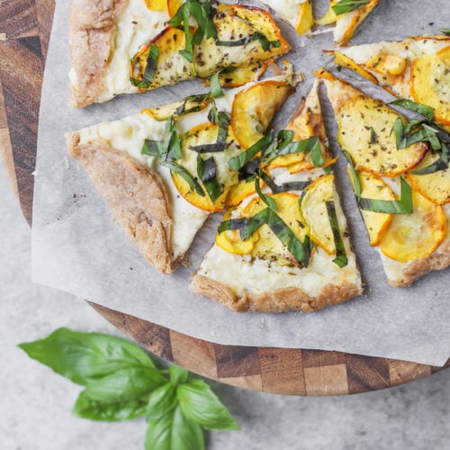 Cheesy Zucchini Tart 14 Six Slices Of Veggie Pizza With Yellow Squash, Basil, And Cheese On Parchment Atop A Wooden Board—Reminiscent Of A Cheesy Zucchini Tart. Fresh Basil Leaves And A Bowl Of Red Pepper Flakes Are Nearby, With A Bowl Of Yellow Squash At The Bottom Right.