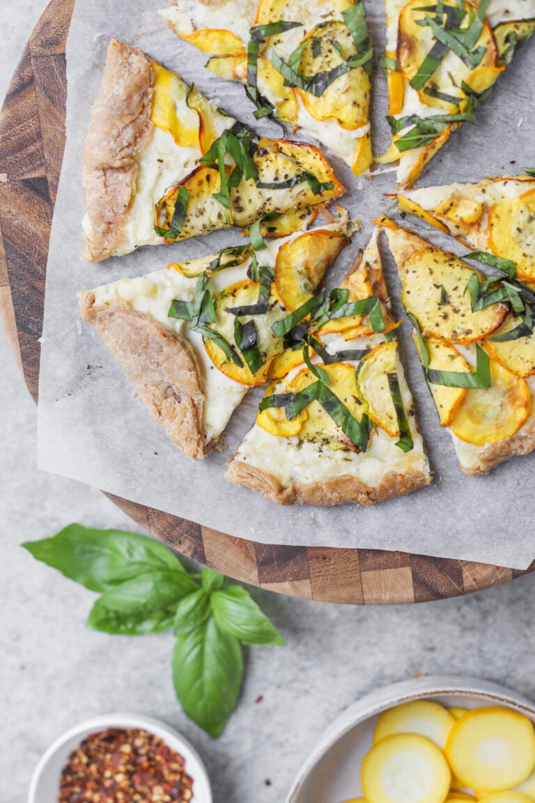 Six Slices Of Veggie Pizza With Yellow Squash, Basil, And Cheese On Parchment Atop A Wooden Board—Reminiscent Of A Cheesy Zucchini Tart. Fresh Basil Leaves And A Bowl Of Red Pepper Flakes Are Nearby, With A Bowl Of Yellow Squash At The Bottom Right.