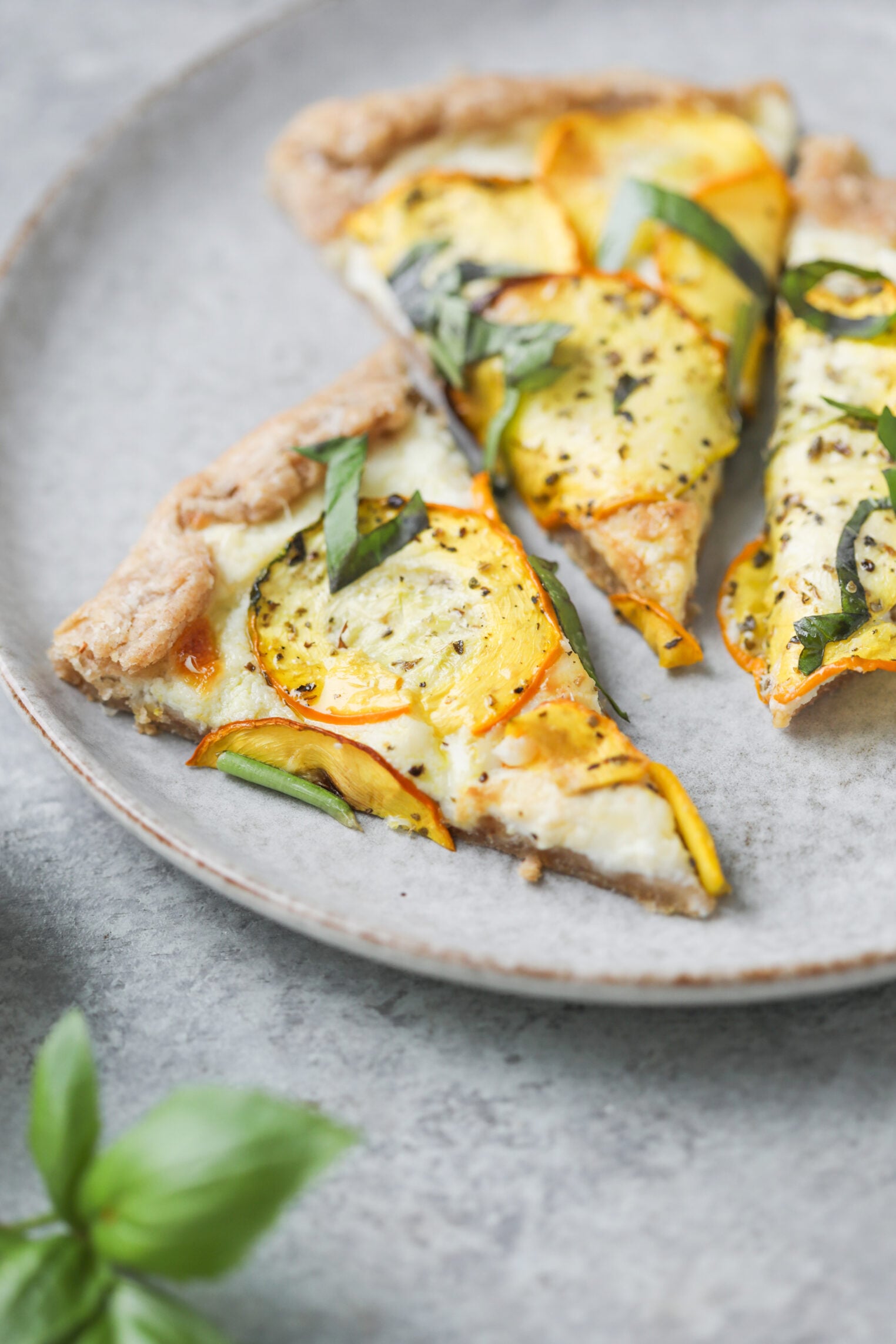 Cheesy Zucchini Tart 1 Two Slices Of Cheesy Zucchini Tart, Topped With Yellow And Green Zucchini, Fresh Basil, And Melted Cheese, Sit On A Gray Ceramic Plate.