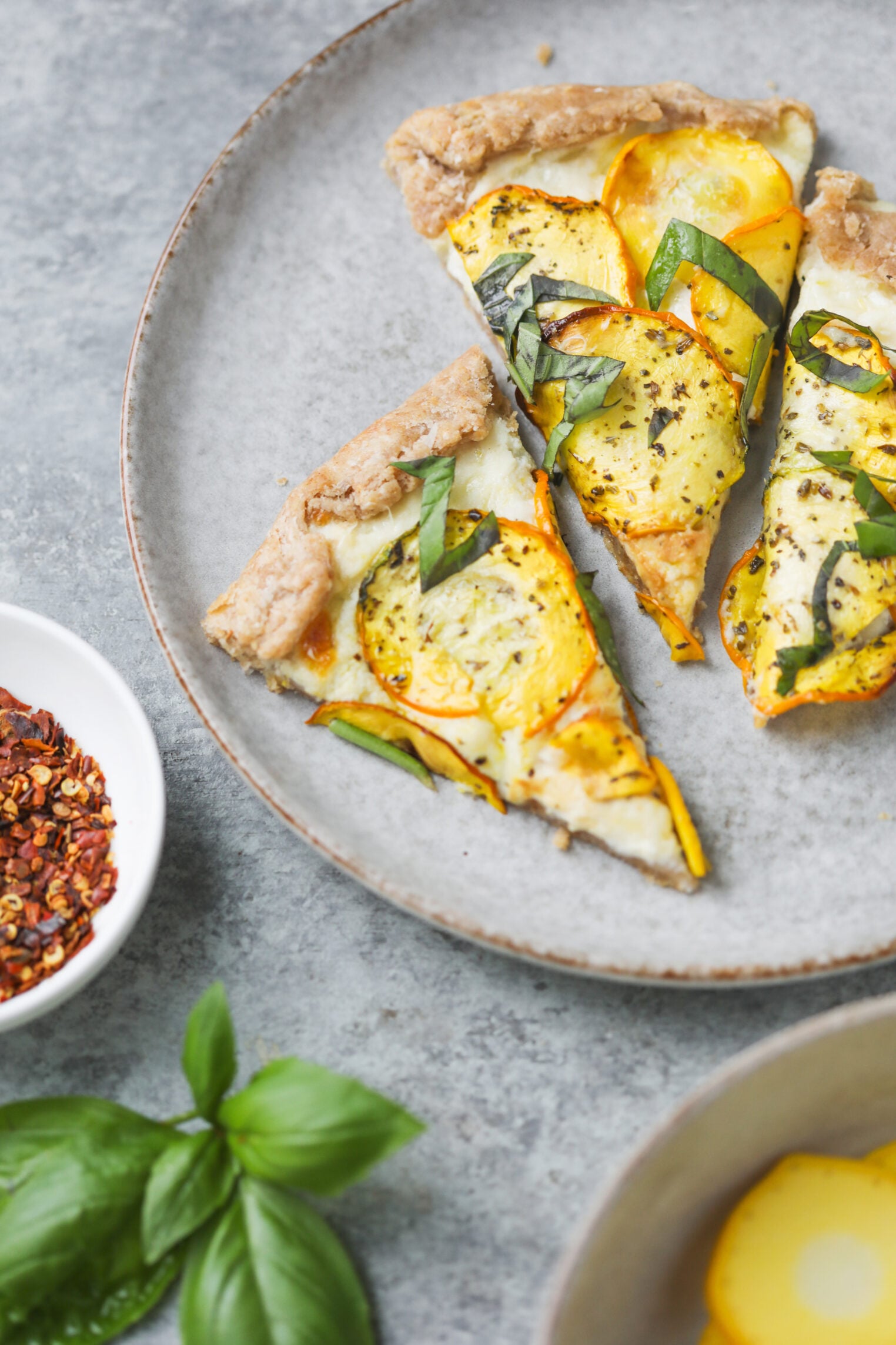 Cheesy Zucchini Tart 3 Three Slices Of Veggie Pizza With Yellow Squash, Basil, And Herbs On A Gray Plate—Reminiscent Of A Cheesy Zucchini Tart. Fresh Basil Leaves And A Bowl Of Red Pepper Flakes Sit Nearby.