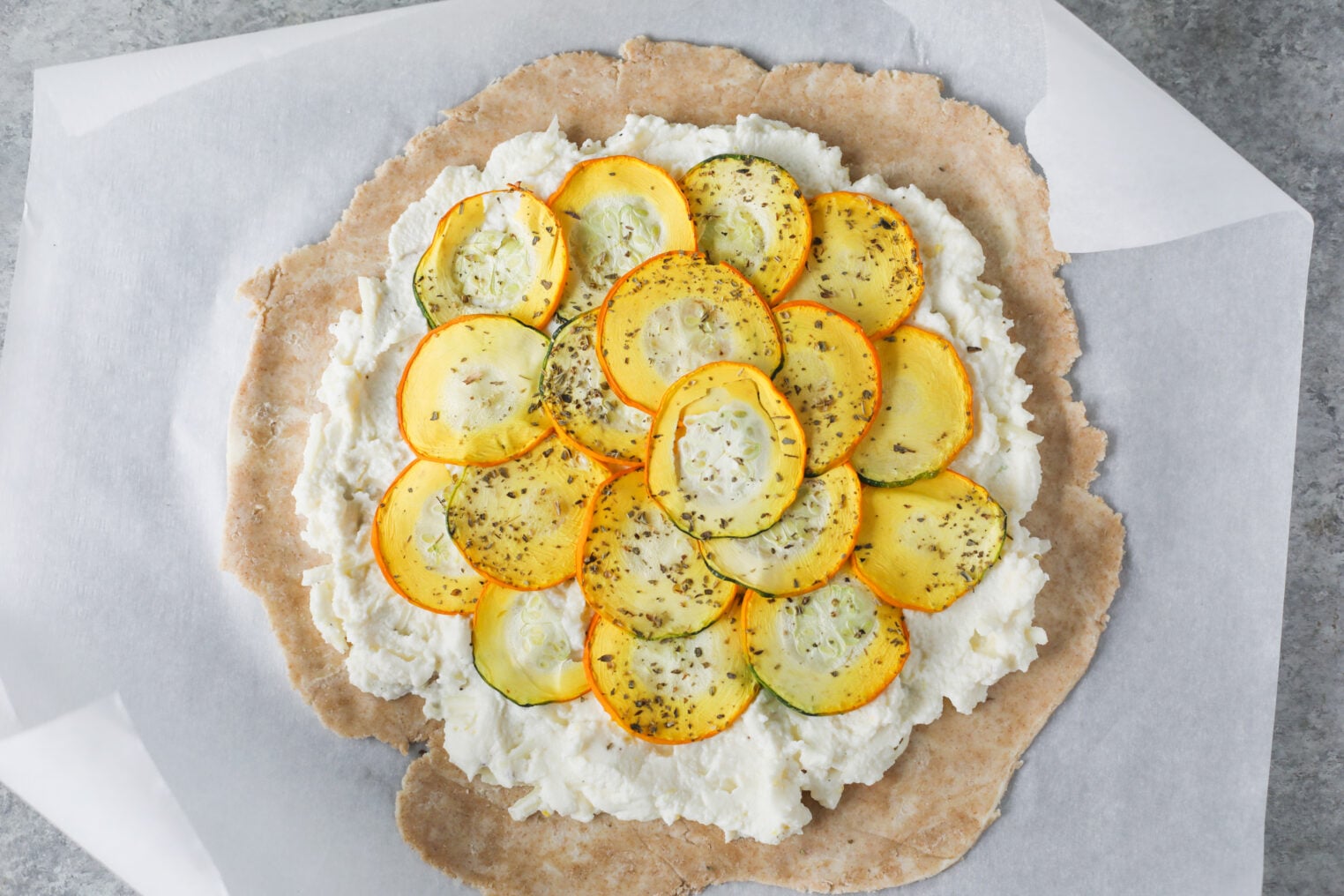 Cheesy Zucchini Tart 10 A Round Zucchini Tart Topped With White Cheese And Overlapping Yellow And Green Zucchini Slices, Sprinkled With Herbs, Sits On Parchment Paper On A Gray Surface.