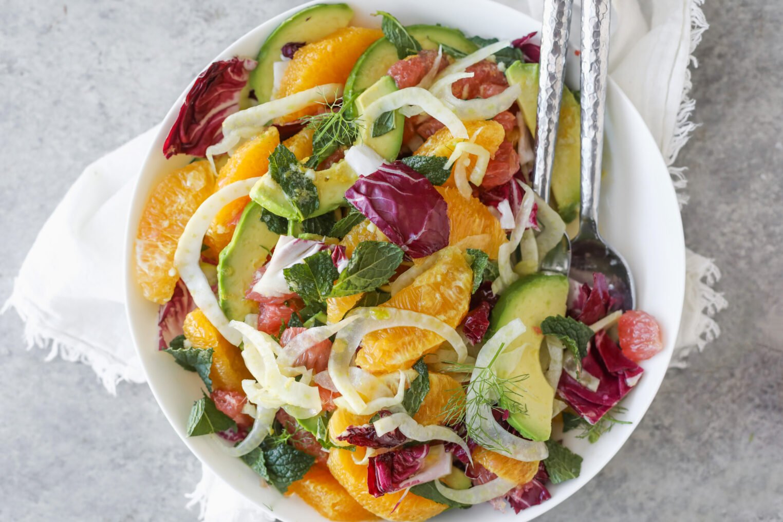 A Fresh Orange And Fennel Salad Is Served In A White Bowl With Avocado, Radicchio, Mint Leaves, And Grapefruit Segments, Set On A Gray Surface Alongside A White Napkin And Serving Utensils.