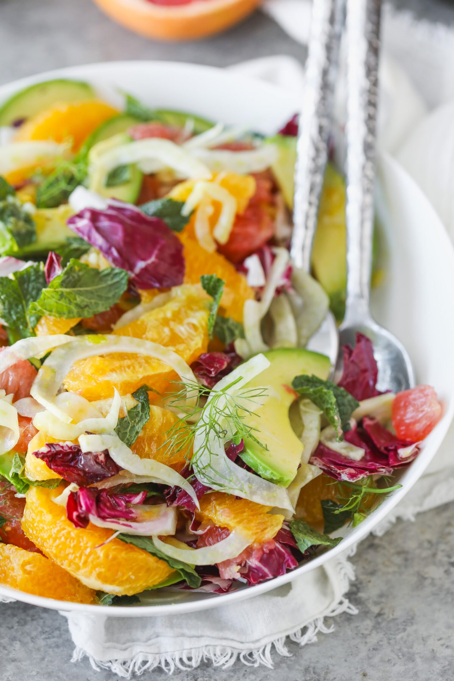 A Vibrant Orange And Fennel Salad With Avocado, Radicchio, Fresh Mint, And Dill, Served In A White Bowl With Metal Salad Servers On A White Napkin.