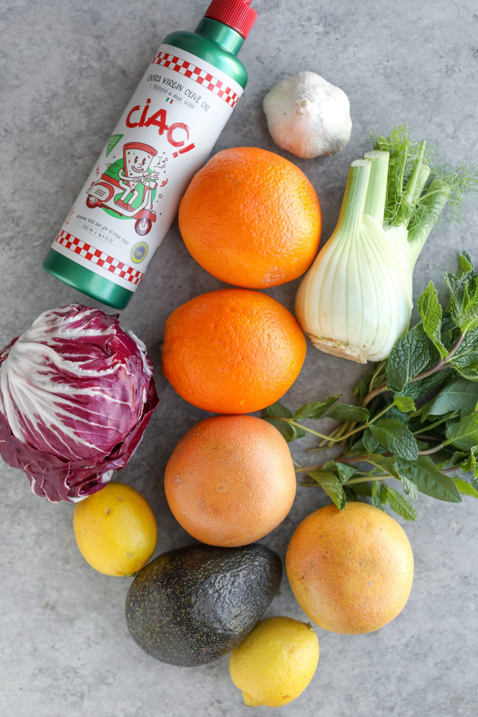 A Bottle Of Olive Oil, A Garlic Bulb, Grapefruit, Oranges, A Fennel Bulb, Mint, Lemon, Avocado, And Radicchio Are Artfully Arranged On A Gray Surface—Perfect Ingredients For A Refreshing Fennel Salad.