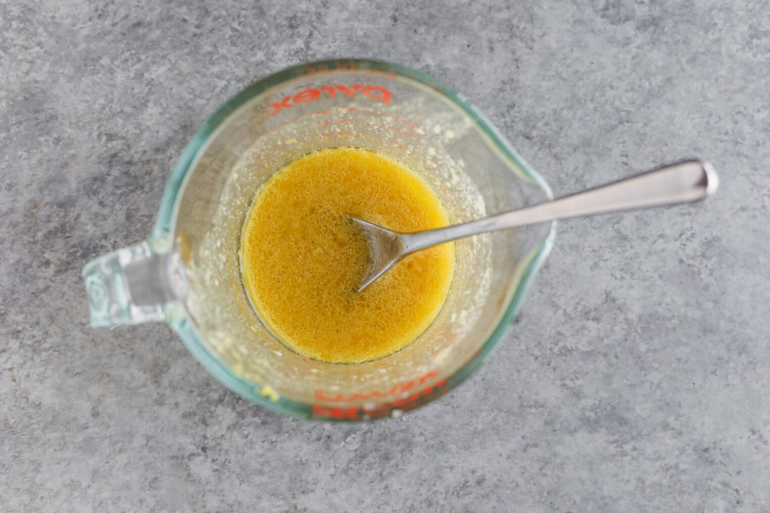 A Metal Spoon Rests In A Glass Measuring Cup Filled With A Yellow Liquid Mixture For An Orange And Fennel Salad, Viewed From Above On A Gray Countertop.