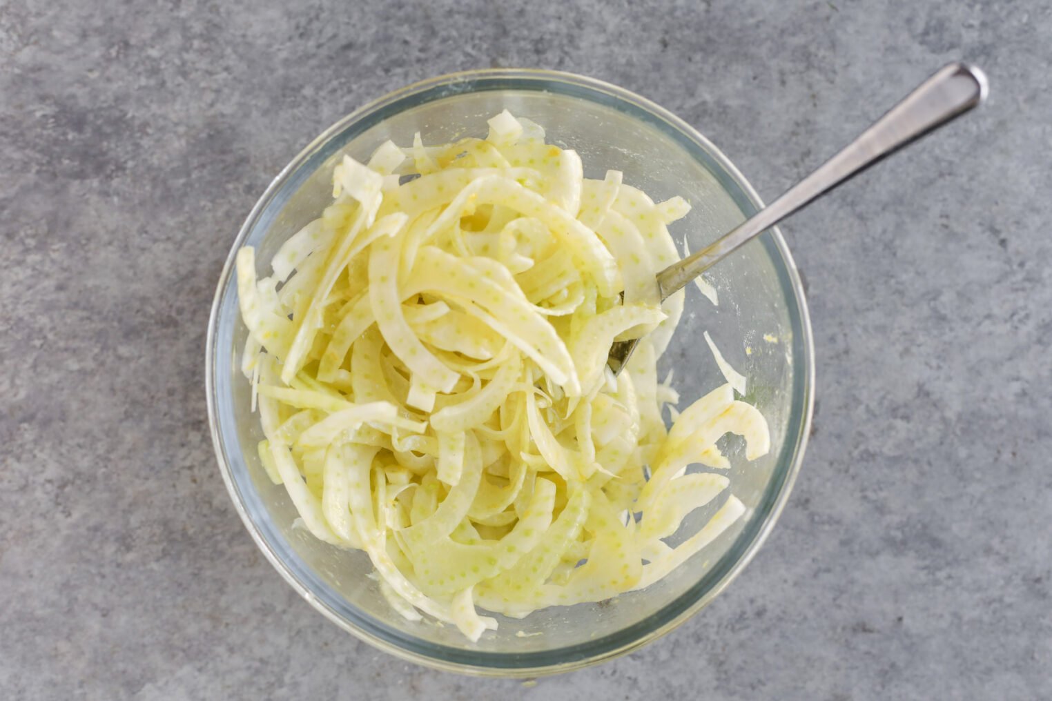 A Glass Bowl Filled With Thinly Sliced Orange And Fennel Salad, Lightly Dressed, With A Metal Fork Resting Inside, Placed On A Gray Textured Surface.