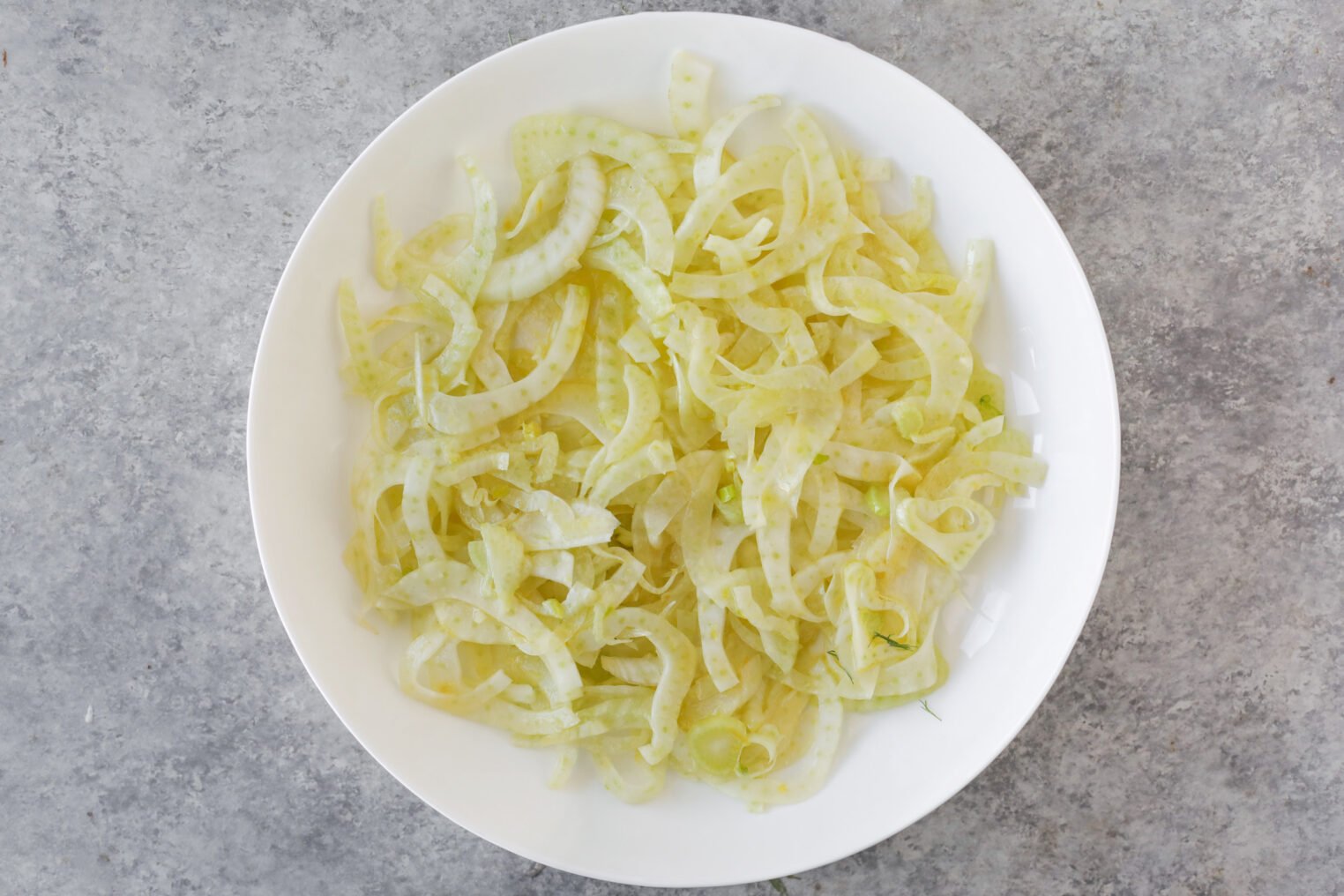 A White Plate Filled With Thinly Sliced Fennel Sits On A Gray, Textured Surface. The Fresh, Pale Green Fennel Hints At A Refreshing Orange And Fennel Salad—Crisp And Vibrant, Ready To Serve.