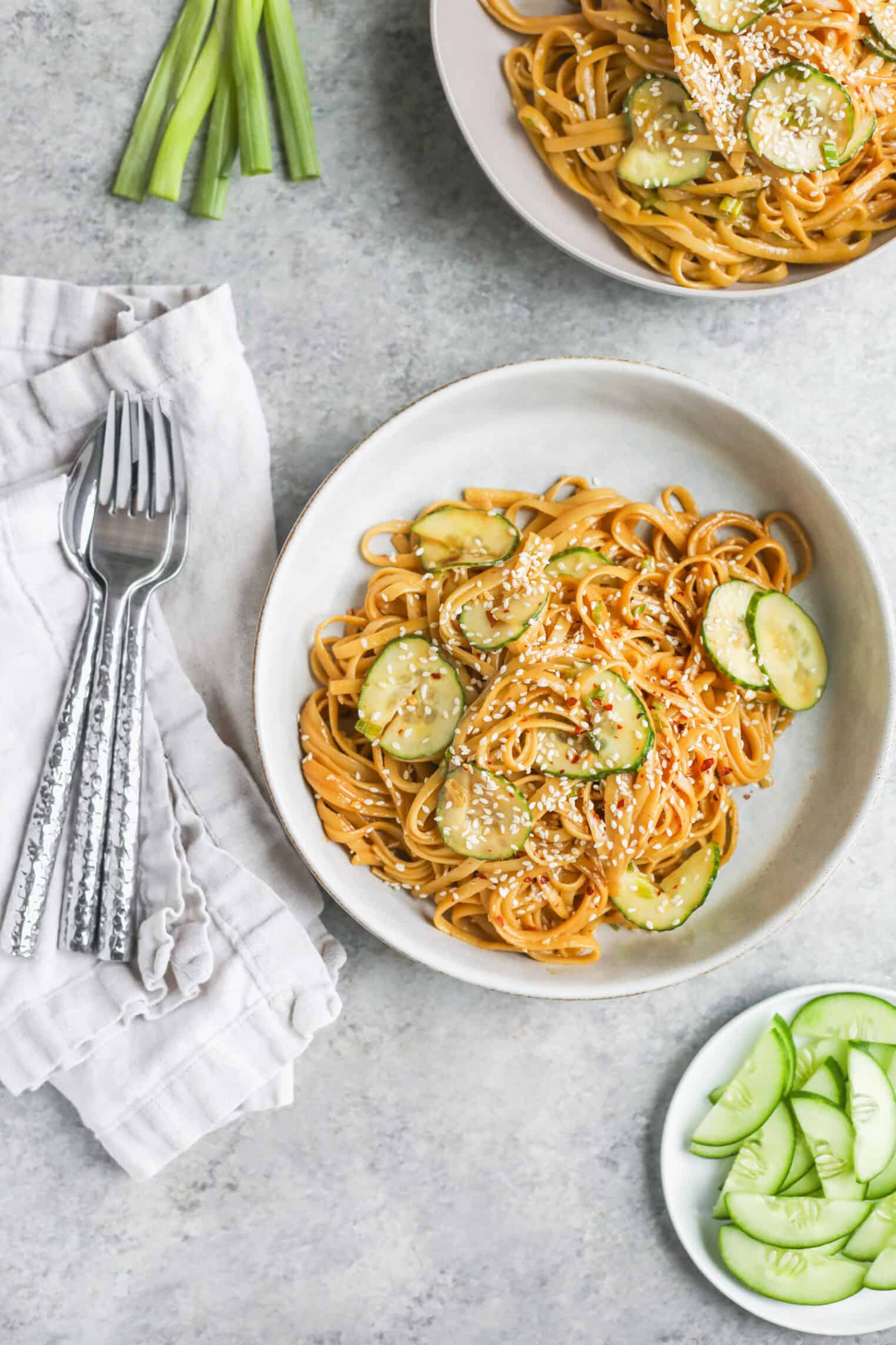 Cold Sesame Noodles With Cucumbers 9 Two Bowls Of Cold Sesame Noodles With Sliced Cucumbers And Sesame Seeds Sit On A Gray Surface. A Small Plate Of Extra Cucumber Slices, A White Napkin, And Three Forks Are Beside The Bowls. Green Onions Are Visible At The Top Left.