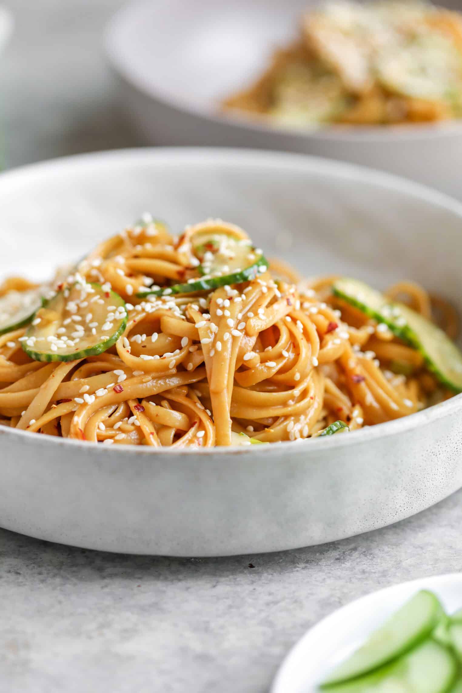 Cold Sesame Noodles With Cucumbers 2 A White Bowl Filled With Cold Sesame Noodles Tossed In A Savory Sauce, Garnished With Sesame Seeds And Sliced Cucumbers. In The Background, Another Bowl Of Food And A Plate With Cucumber Slices Are Visible.
