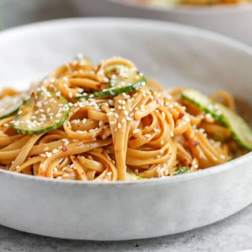 Cold Sesame Noodles With Cucumbers 11 A White Bowl Filled With Cold Sesame Noodles Tossed In A Savory Sauce, Garnished With Sesame Seeds And Sliced Cucumbers. In The Background, Another Bowl Of Food And A Plate With Cucumber Slices Are Visible.