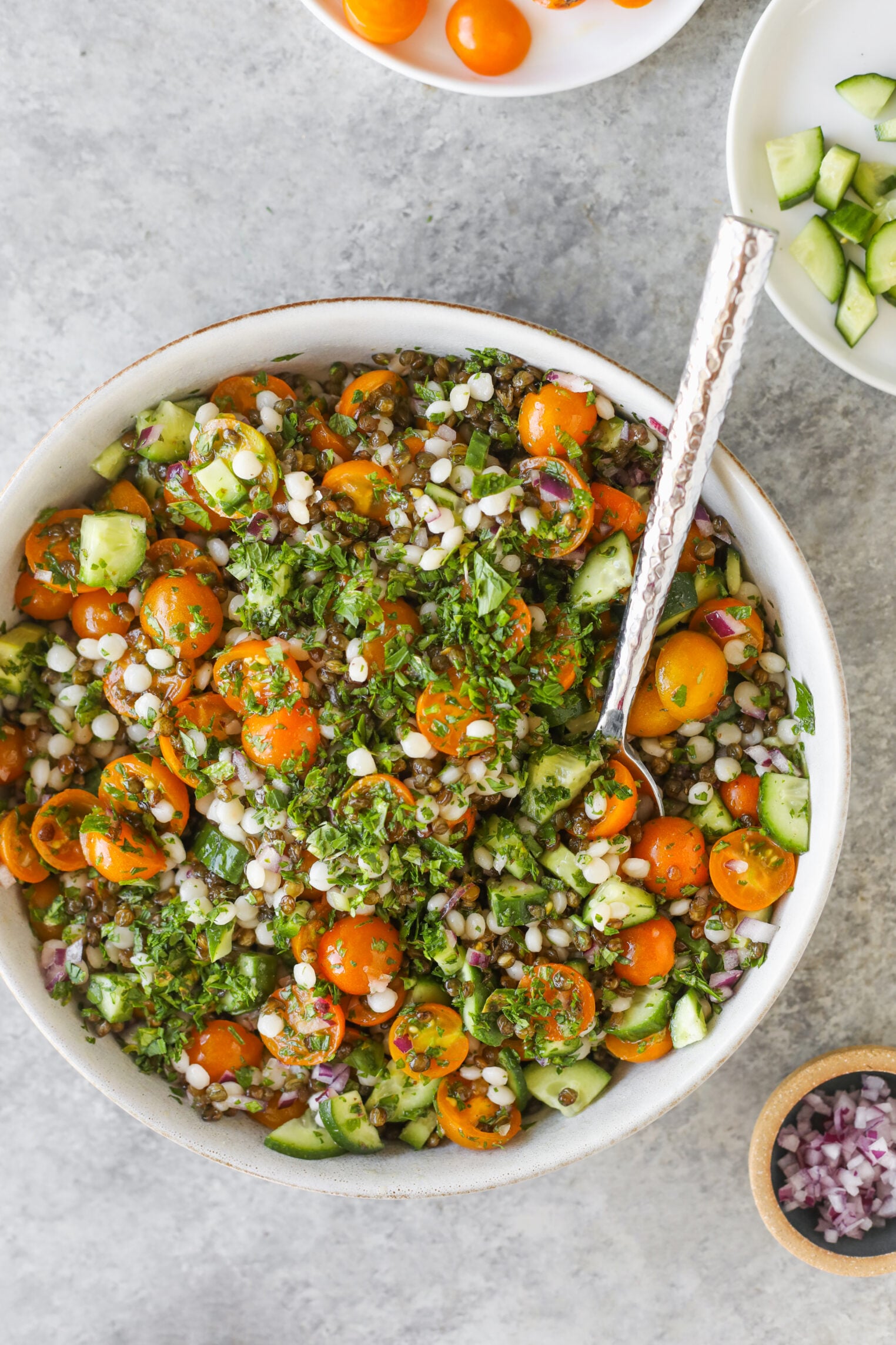A Bowl Of Salad With Cherry Tomatoes, Cucumbers, Chopped Herbs, Pearl Couscous, And Red Onions, With A Spoon Inside—Reminiscent Of A Refreshing Lentil Tabbouleh. Small Bowls Of Diced Cucumber And Tomatoes Are Nearby.