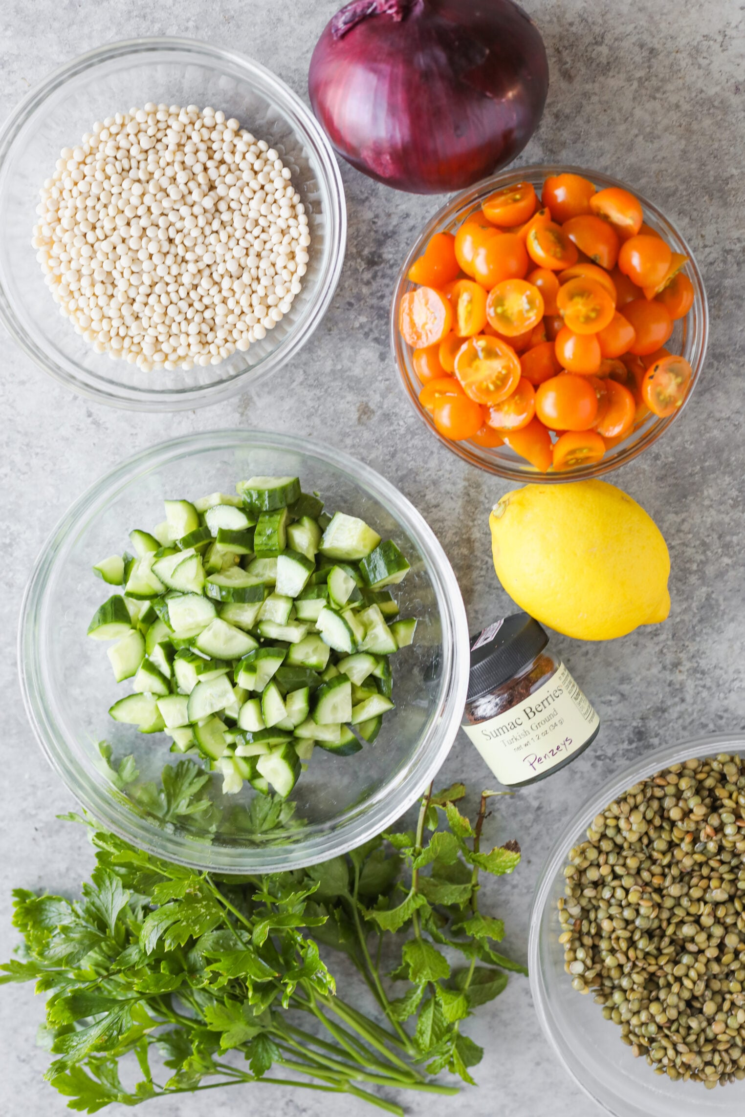 Bowls Of Uncooked Couscous, Halved Orange Cherry Tomatoes, Chopped Cucumber, Green Lentils, A Red Onion, A Lemon, Fresh Parsley, And A Jar Of Spice Are Arranged On A Gray Surface—Perfect Ingredients For Creating Vibrant Lentil Tabbouleh.