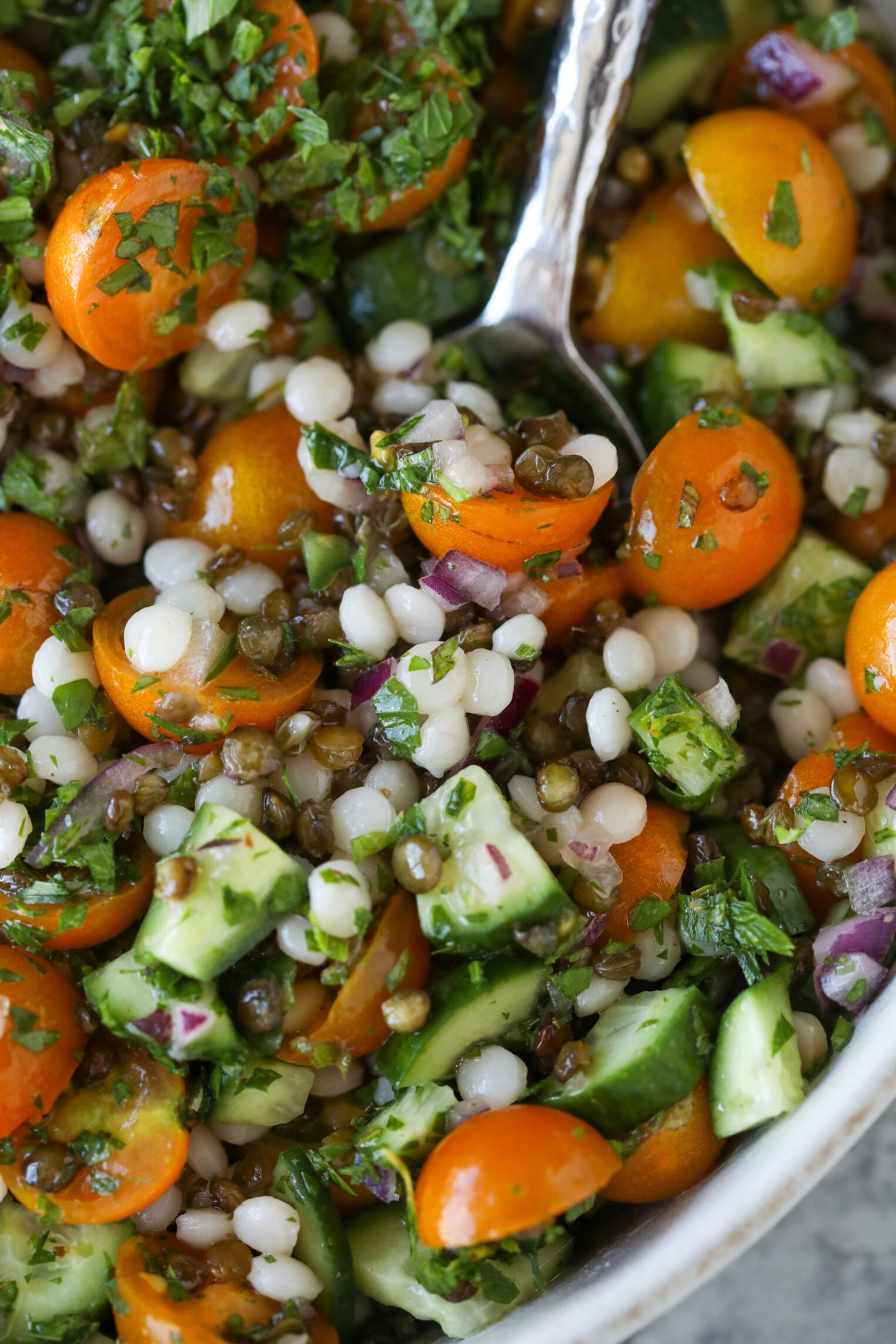 Close-Up Of A Fresh Salad Inspired By Lentil Tabbouleh, Featuring Pearl Couscous, Cherry Tomatoes, Diced Cucumbers, Red Onion, Lentils, And Chopped Herbs, All Mixed Together In A Bowl With A Spoon.