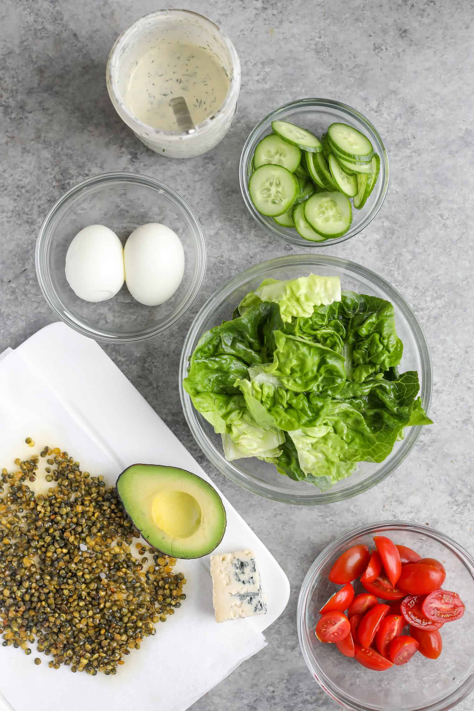 Vegetarian Cobb Salad With Crispy Lentils 3 A Flat Lay Of Salad Ingredients For A Fresh Cobb Salad: Leafy Lettuce, Sliced Cucumbers, Grape Tomatoes, Halved Avocado, Boiled Eggs, Blue Cheese, A Jar Of Dressing, And Crunchy Seeds Rest On A White Napkin.