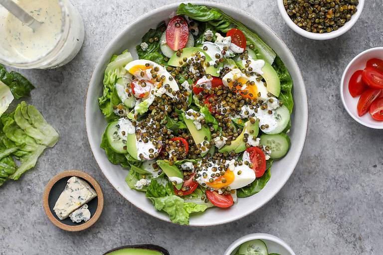 A Vibrant Cobb Salad Bowl With Lettuce, Cherry Tomatoes, Avocado Slices, Hard-Boiled Egg Wedges, Crispy Lentil Topping, And Creamy Dressing. Surrounding The Bowl Are More Tomatoes, Blue Cheese, Avocado, Lettuce, And A Jar Of Dressing.