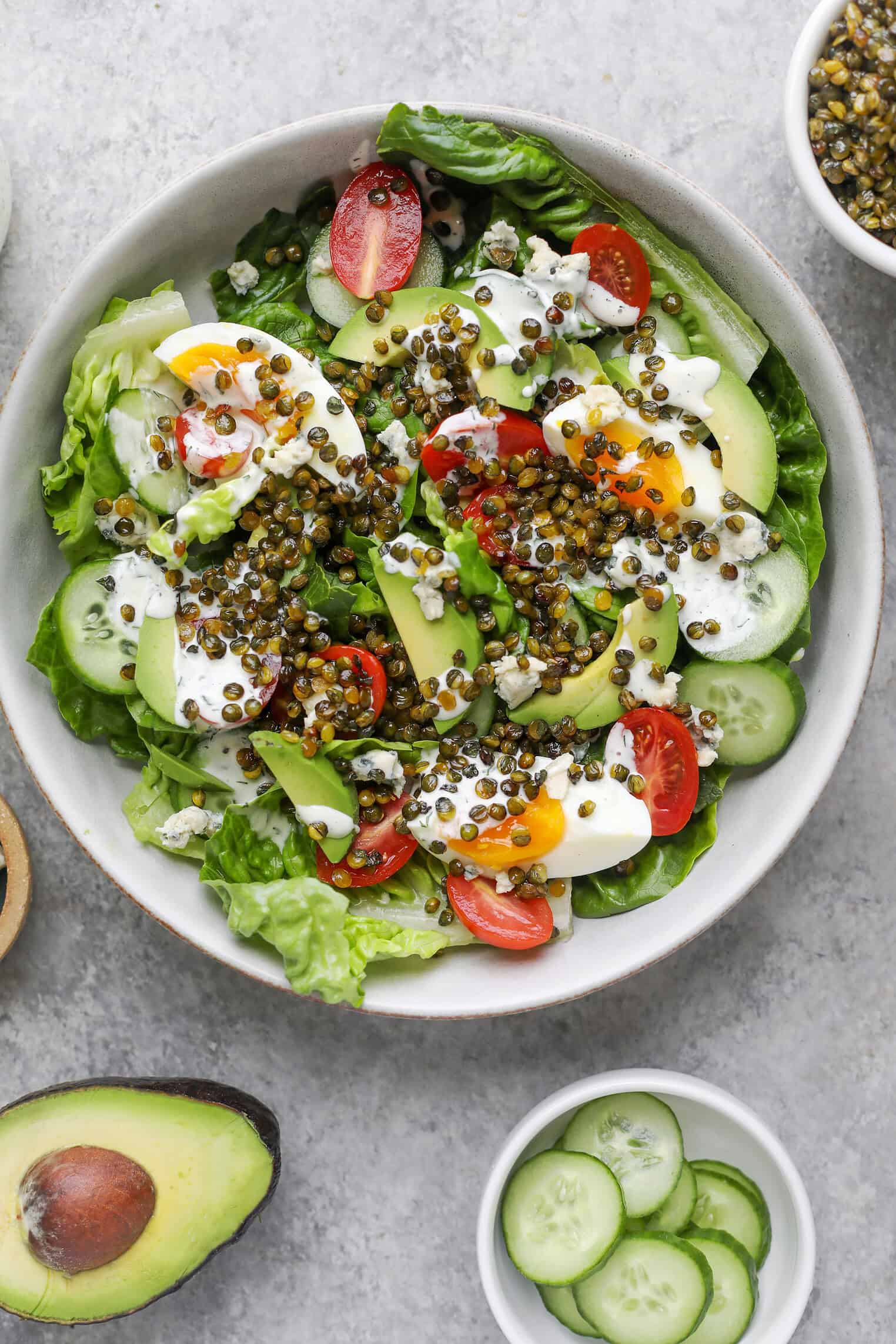 A Fresh Lentil Salad In A Bowl Featuring Lettuce, Cucumber Slices, Cherry Tomato Halves, Avocado, Boiled Egg Slices, Blue Cheese Crumbles, Crispy Lentils, And A Drizzle Of Creamy Dressing. Avocado And Cucumbers Are Nearby.