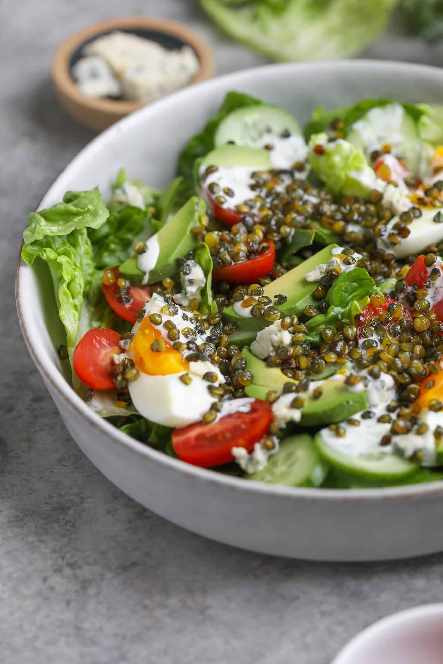 Vegetarian Cobb Salad With Crispy Lentils 8 A Fresh Lentil Cobb Salad In A White Bowl Features Cherry Tomatoes, Sliced Cucumber, Avocado, Lettuce, Soft-Boiled Eggs, Lentils, And Creamy Dressing. A Small Dish Of Blue Cheese Sits In The Background.