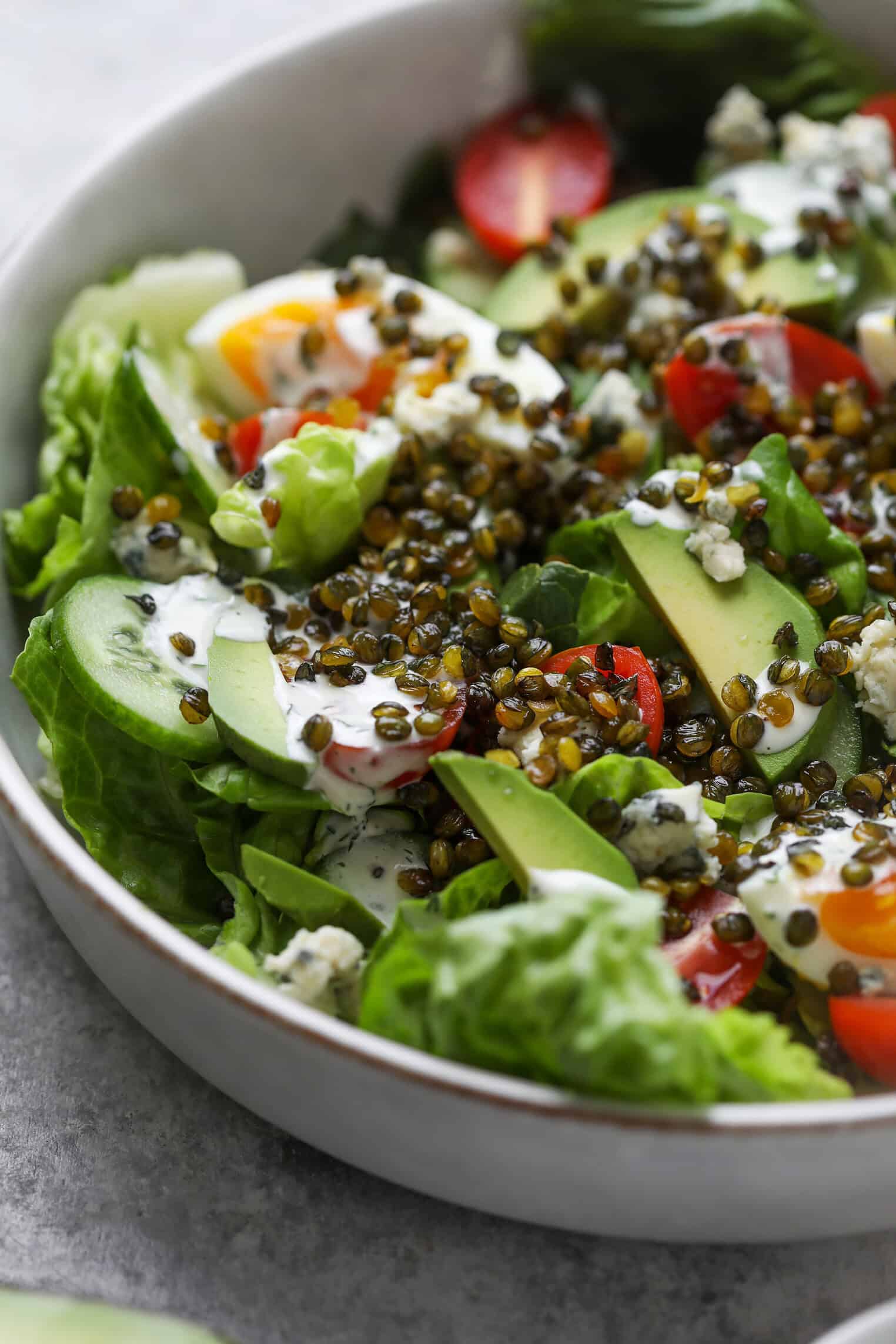 Vegetarian Cobb Salad With Crispy Lentils 2 A Close-Up Of A Fresh Lentil Salad With Lettuce, Sliced Avocado, Cherry Tomatoes, Cucumber, Blue Cheese Crumbles, Creamy Dressing, And Crispy Lentils In A White Bowl.