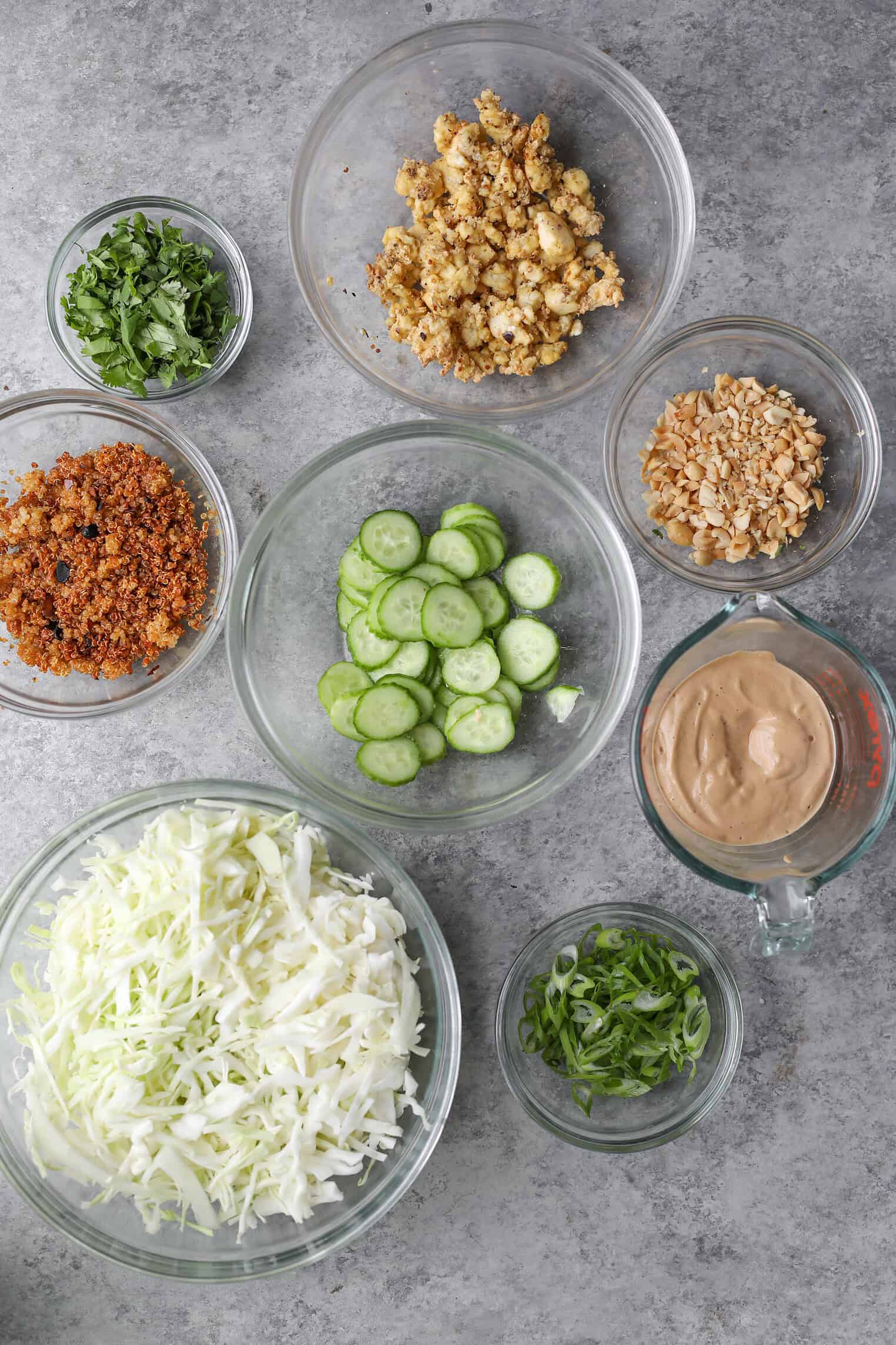 Crispy Quinoa Salad 2 Various Clear Bowls On A Gray Surface Containing Ingredients For A Crispy Quinoa Salad: Shredded Cabbage, Sliced Cucumbers, Chopped Herbs, Crispy Bits, Nuts, Creamy Dressing, Green Onions, And Pieces Of Crispy Tofu Or Tempeh.
