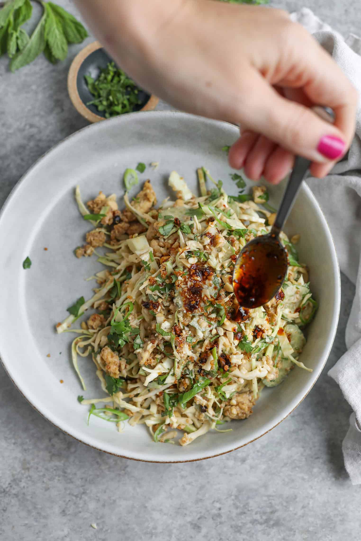A Hand With Pink Nail Polish Drizzles Chili Oil From A Spoon Over A Bowl Of Noodles Topped With Ground Meat, Herbs, And Chopped Greens—Next To A Crispy Quinoa Salad On A Gray Surface.